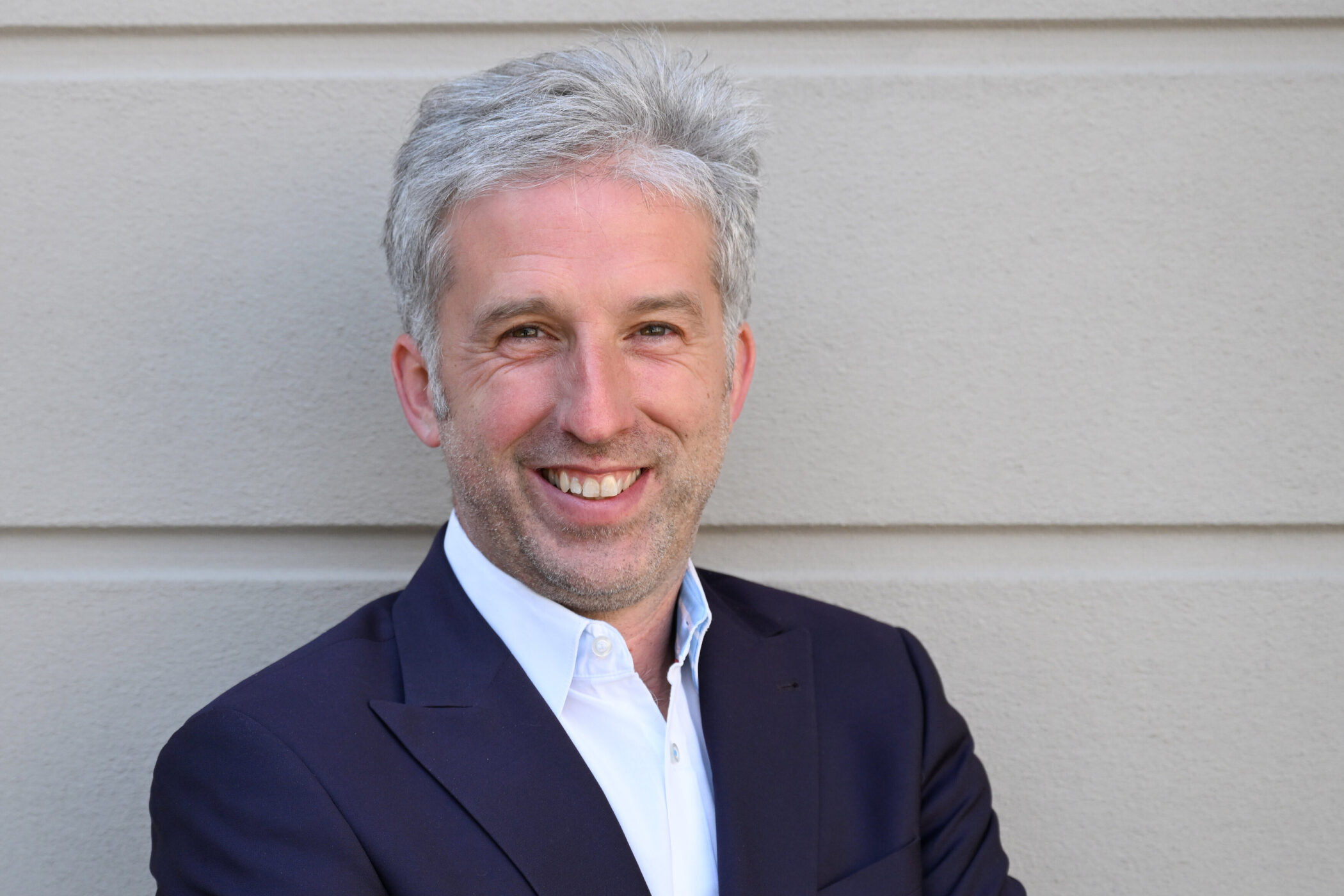 Ein Mann in Anzug lächelt vor einer beigen Wand.