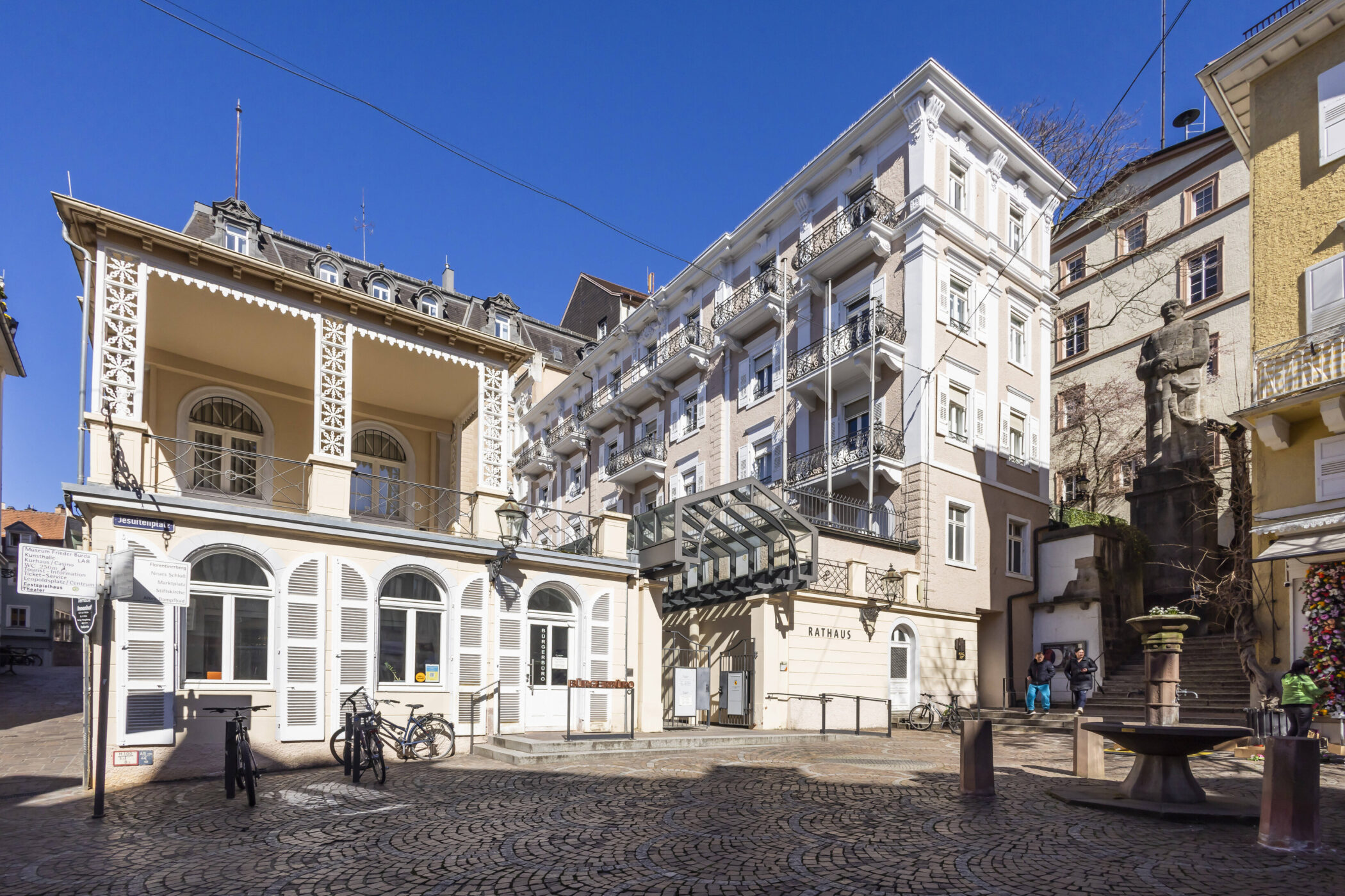 Pflasterplatz mit Brunnen, Statue, Fahrrädern und Gebäuden bei blauem Himmel.