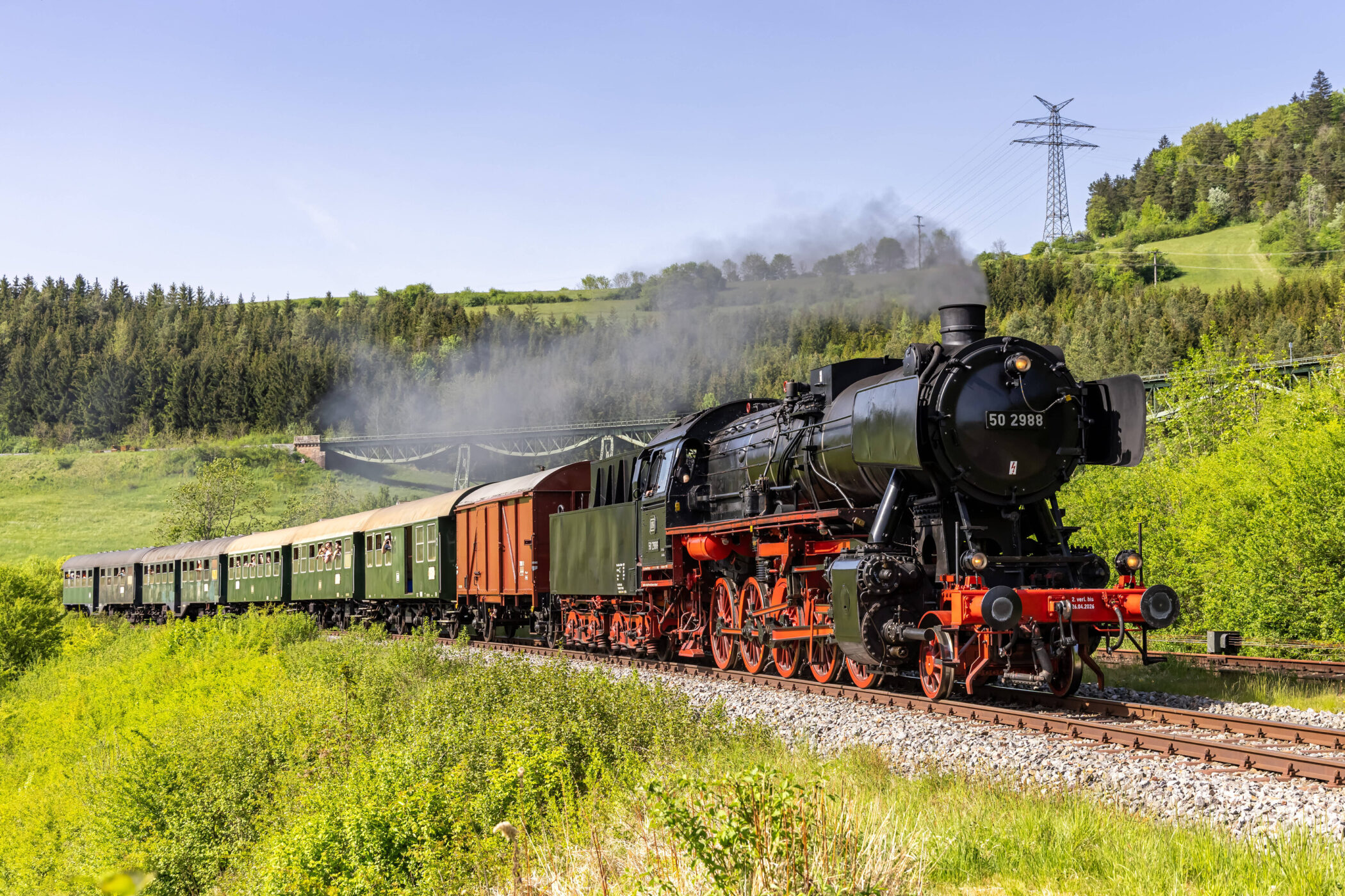 Dampflok mit Wagons fährt durch grüne Landschaft, blauer Himmel im Hintergrund.