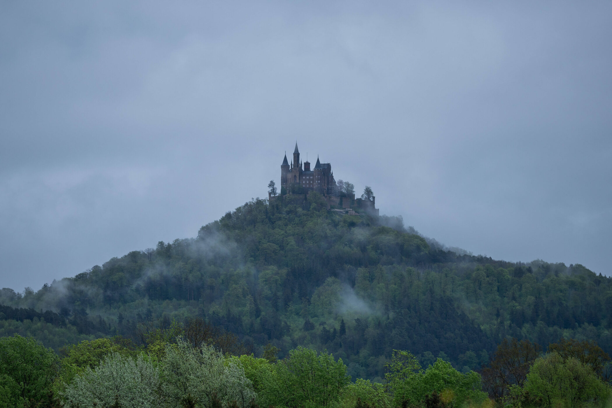 Burg auf bewaldetem Hügel, umgeben von Nebel und Wolken.
