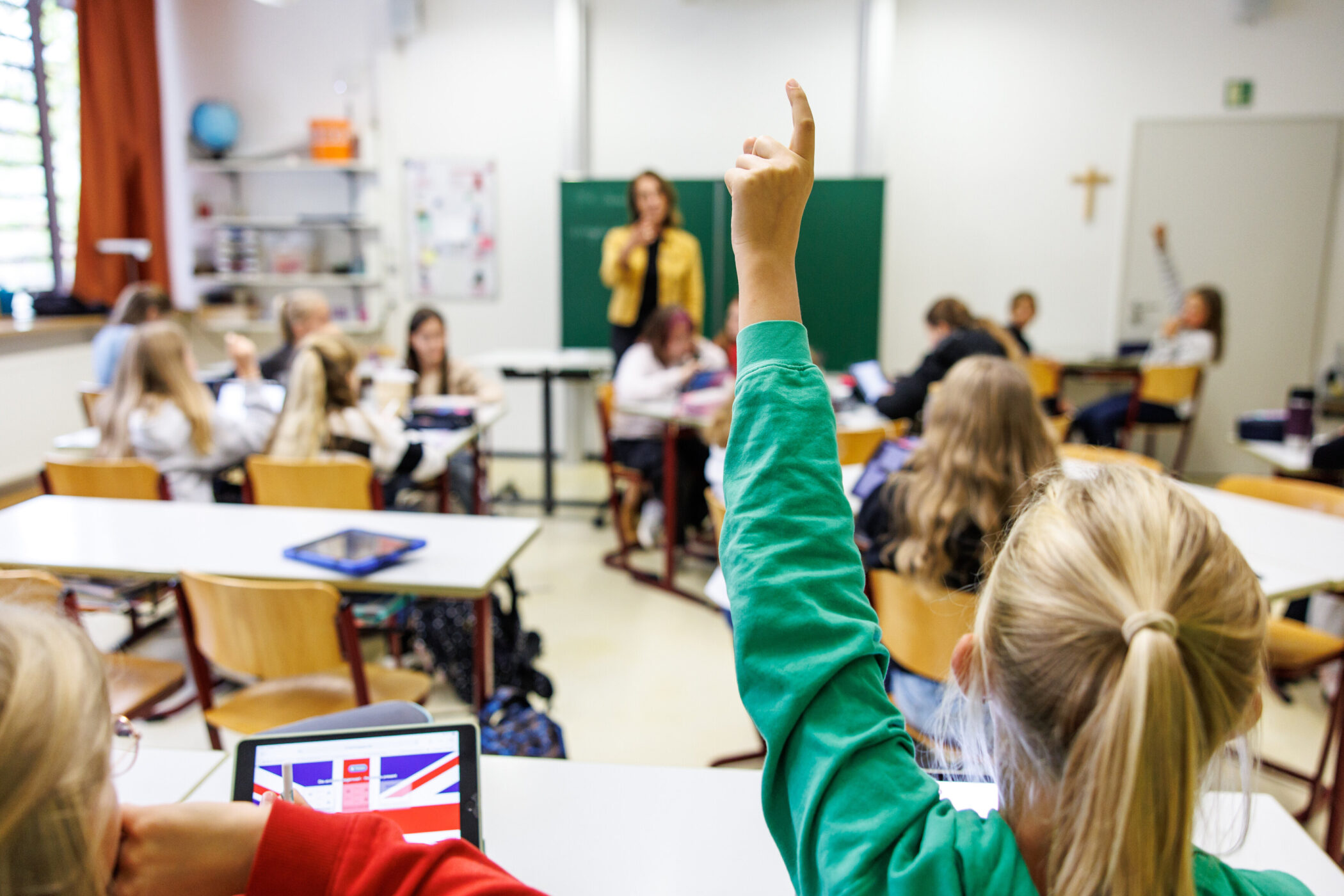 Schüler im Klassenzimmer, eine Person hebt die Hand, Lehrerin vorne.
