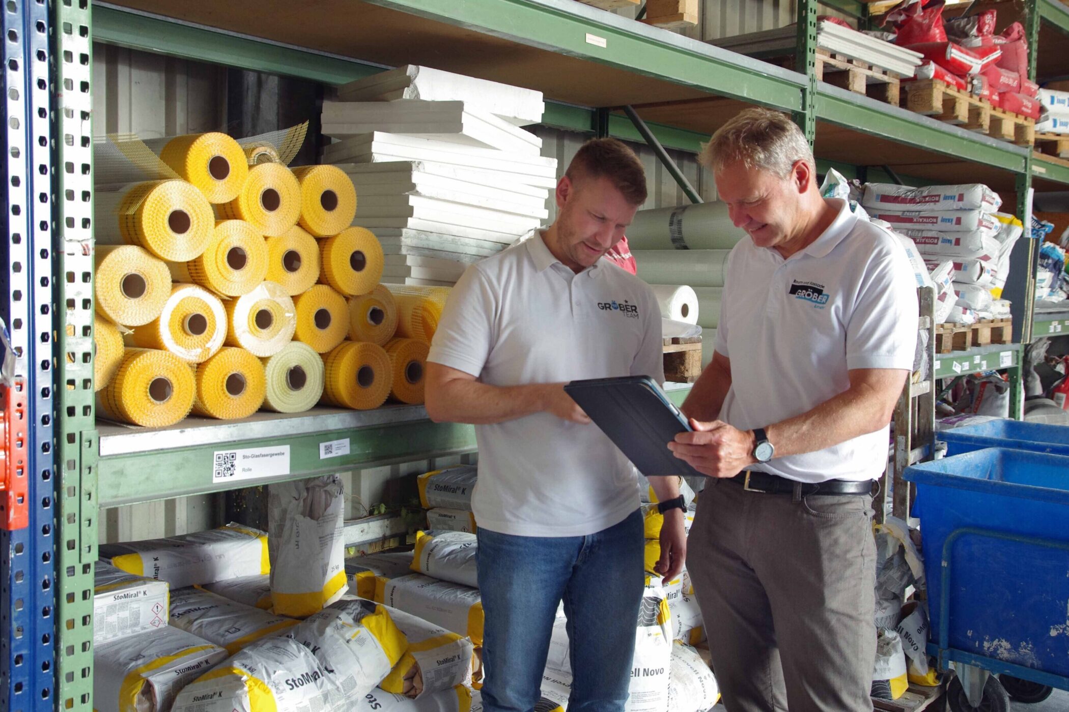 Zwei Männer in einem Lager, betrachten ein Tablet, umgeben von Baumaterialien.
