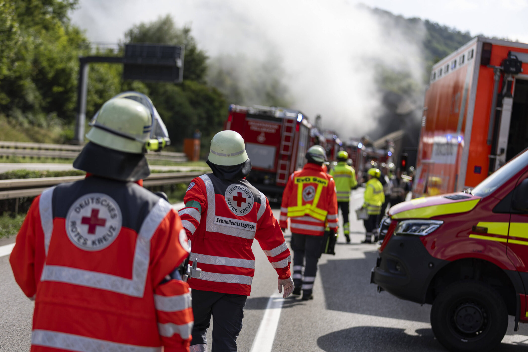 Mehrere Einsatzkräfte in Schutzkleidung auf einer Straße, Rauch im Hintergrund.