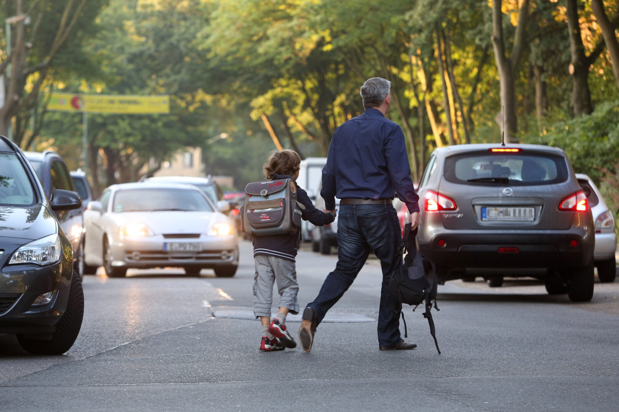 Ein Erwachsener und ein Kind überqueren eine Straße mit Autos im Hintergrund.