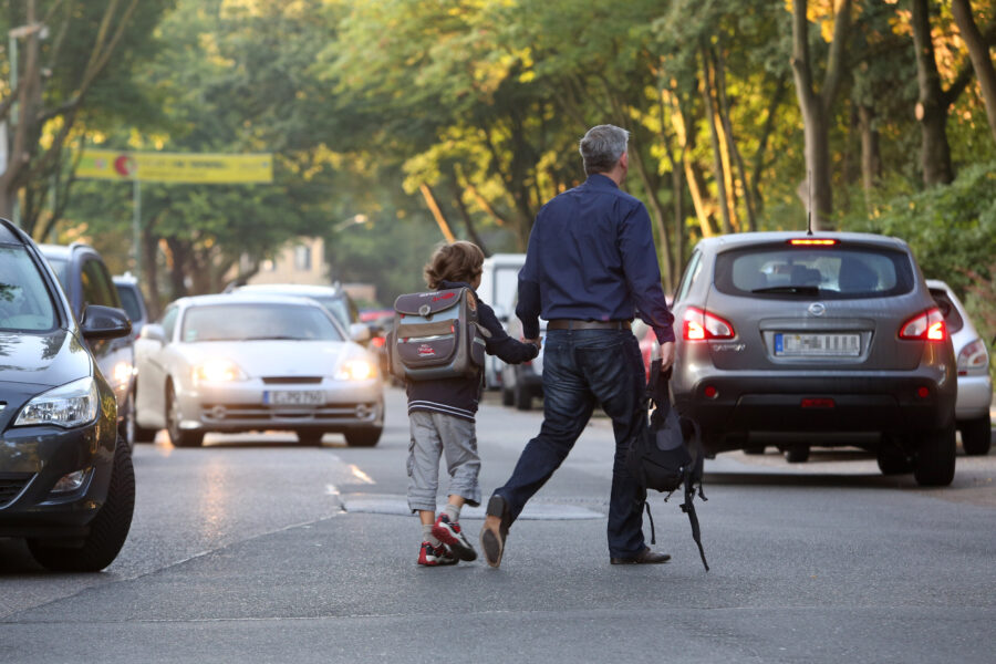 Ein Erwachsener und ein Kind überqueren eine Straße mit Autos im Hintergrund.