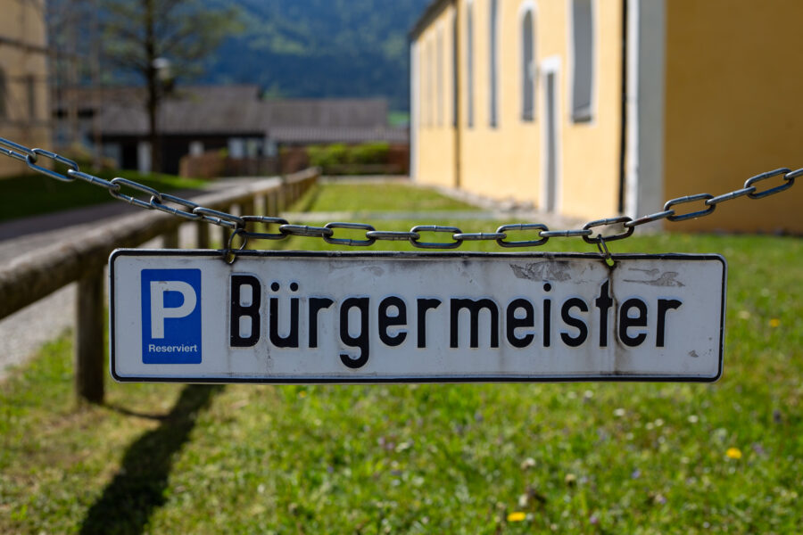 Schild mit Aufschrift "Bürgermeister", Kette, gelbes Gebäude im Hintergrund.
