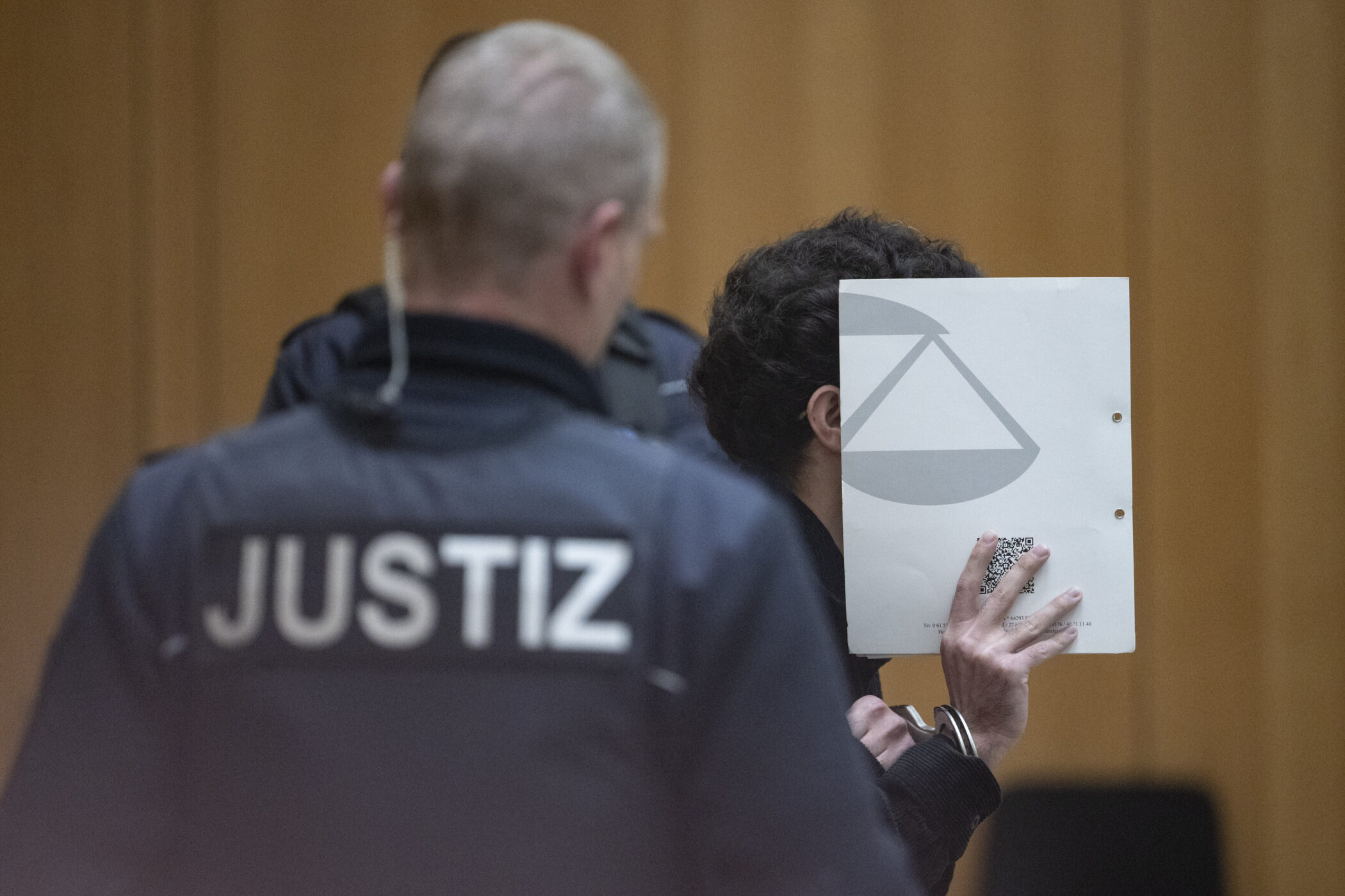 Zwei Personen, eine mit "Justiz"-Aufschrift, andere hält ein Blatt vor das Gesicht.