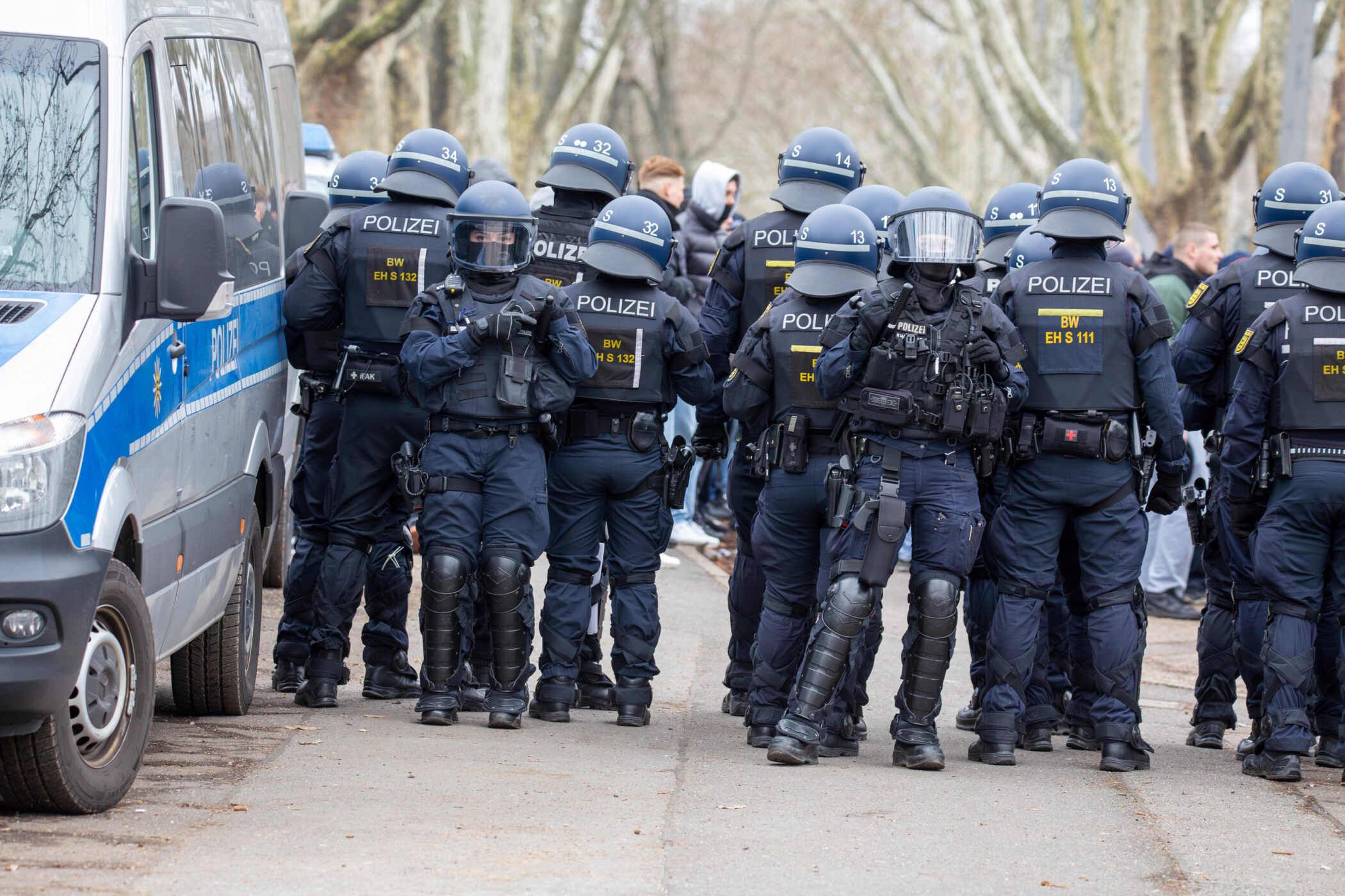 Polizisten in Schutzkleidung stehen vor einem Polizeifahrzeug auf einer Straße.