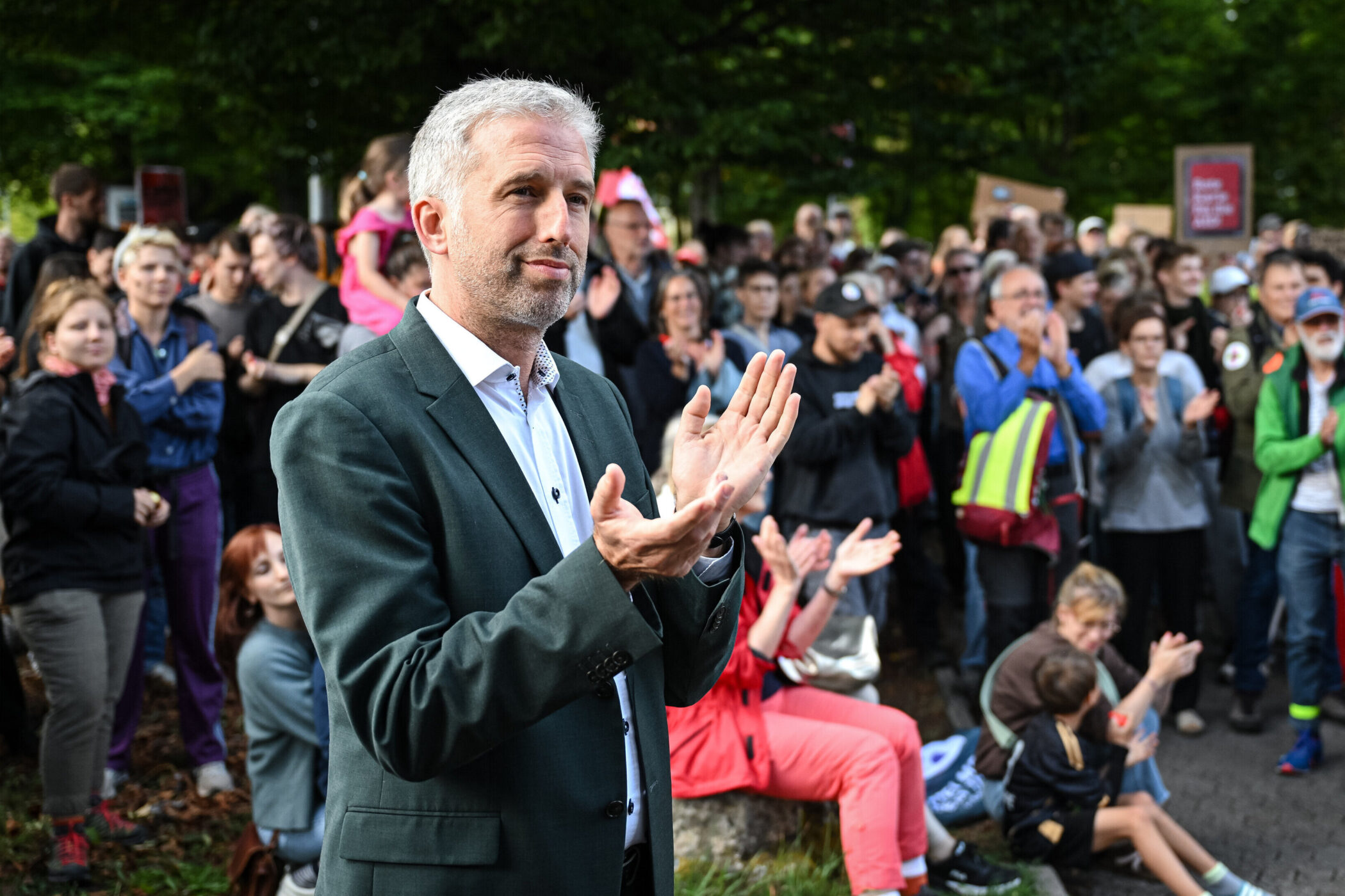 Mann in Anzug klatscht, umgeben von applaudierenden Menschen im Freien.