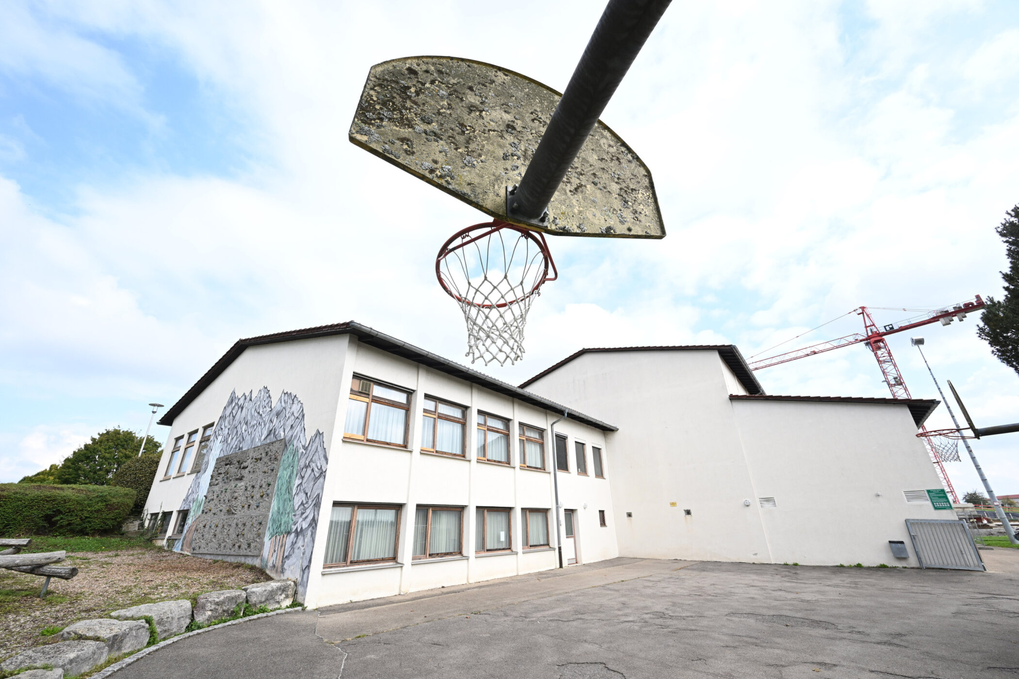 Basketballkorb vor einem weißen Gebäude mit Wandgemälde, Kran im Hintergrund.