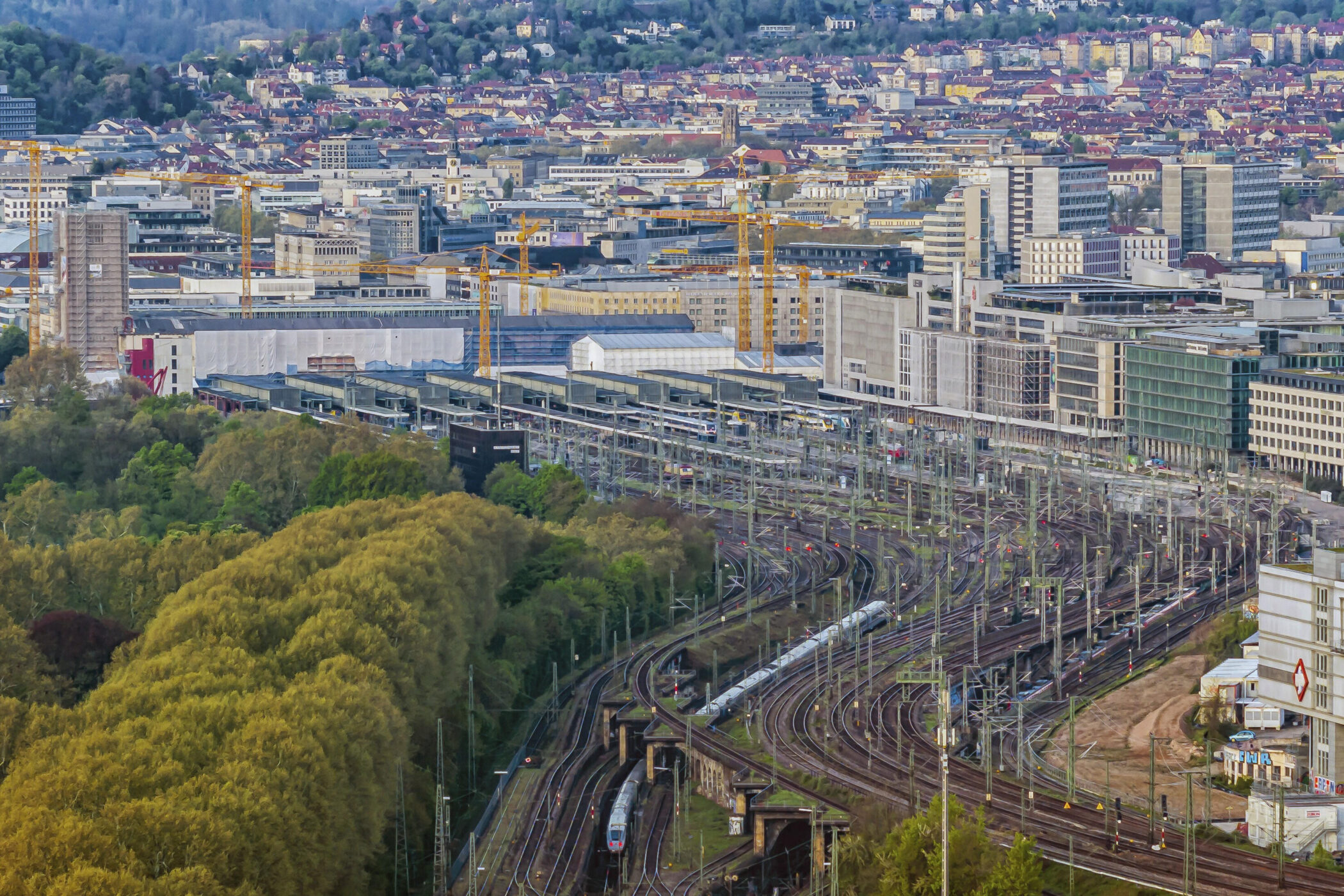 Stadtansicht mit Bahngleisen, Gebäuden und Kränen im Hintergrund.