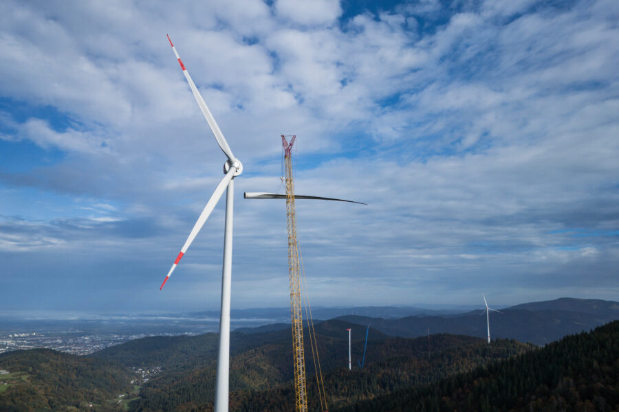 Windrad mit Kran und Landschaft im Hintergrund, bewölkter Himmel.