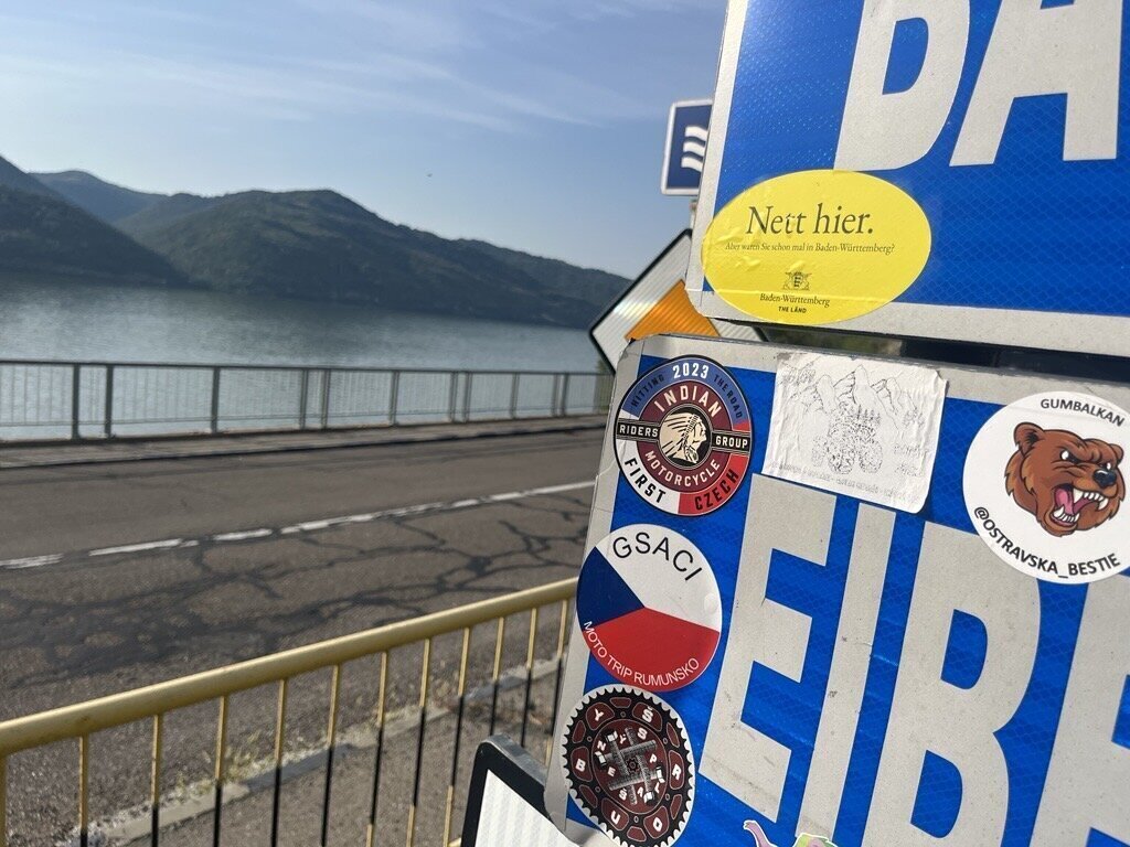 Straßenschild mit Aufklebern, im Hintergrund ein See und Hügel.