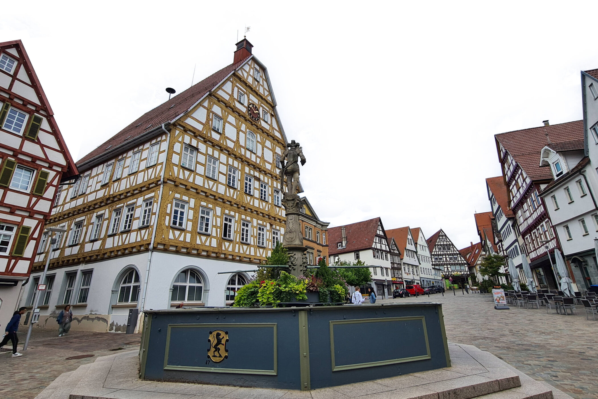 Fachwerkhäuser umgeben einen Platz mit Brunnen und Statue, Menschen gehen vorbei.