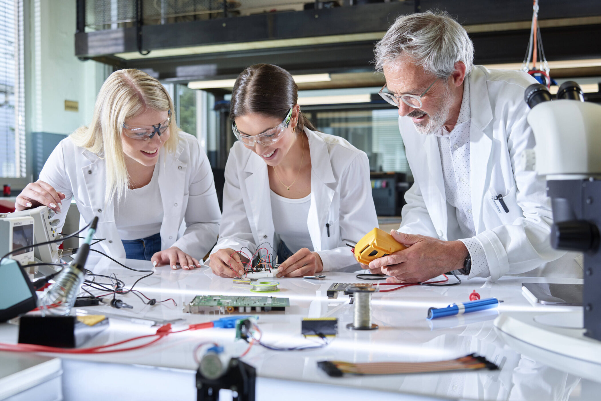 Drei Personen in Laborkitteln arbeiten an elektronischen Geräten.