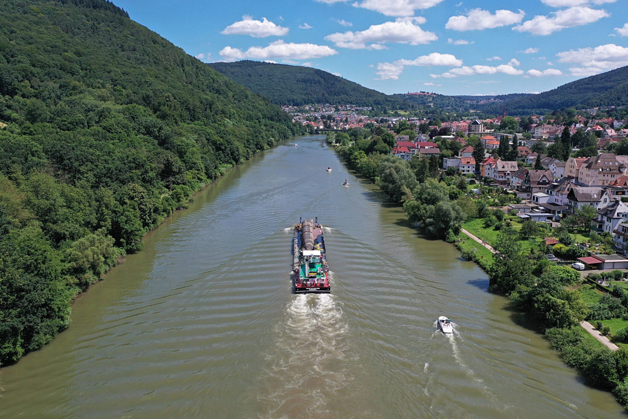 Fluss mit Frachtschiff, umgeben von Wald und Stadt, blauer Himmel mit Wolken.