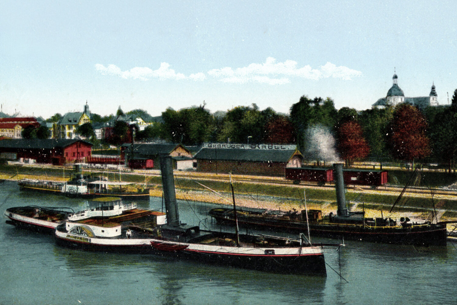 Dampfschiffe auf einem Fluss, im Hintergrund Gebäude und Bäume.