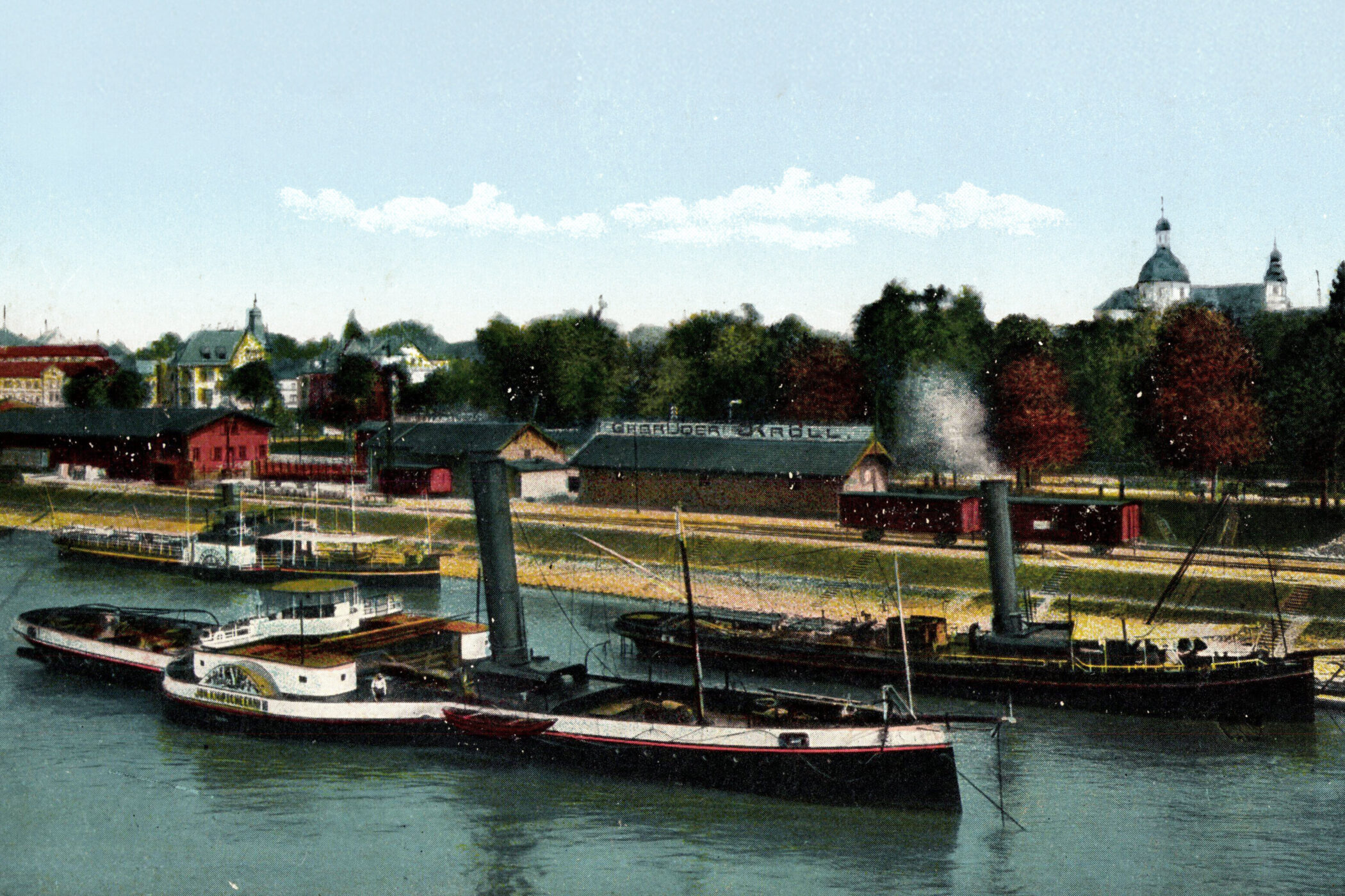 Dampfschiffe auf einem Fluss, im Hintergrund Gebäude und Bäume.