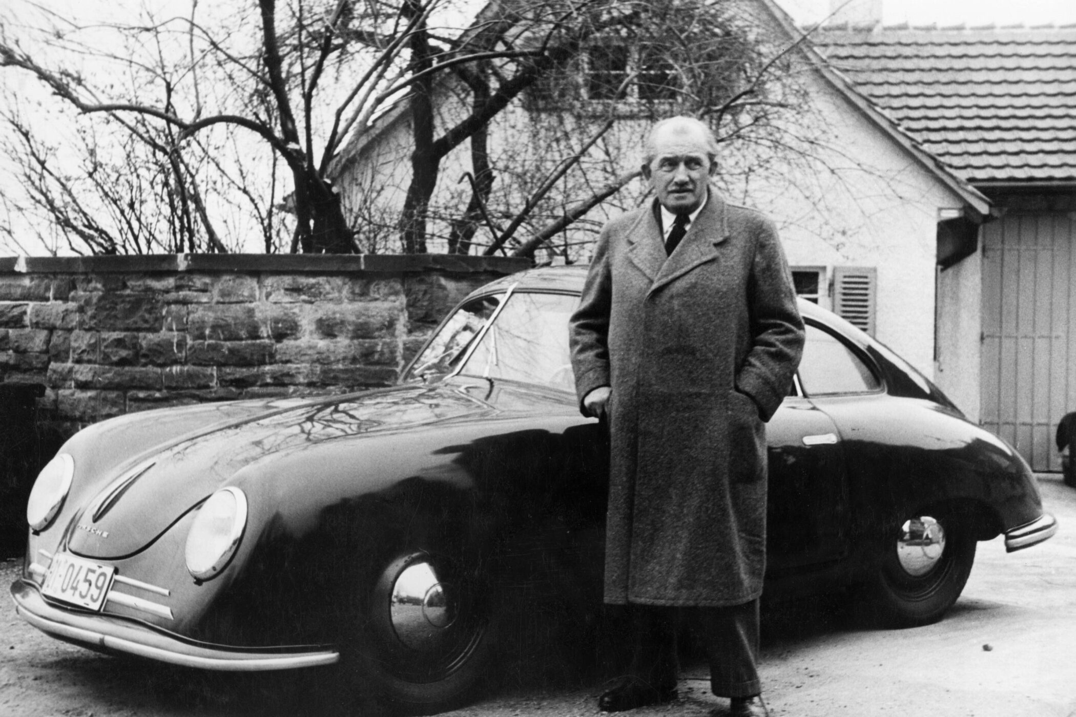 Älterer Mann im Mantel steht neben einem klassischen Auto vor einem Haus.