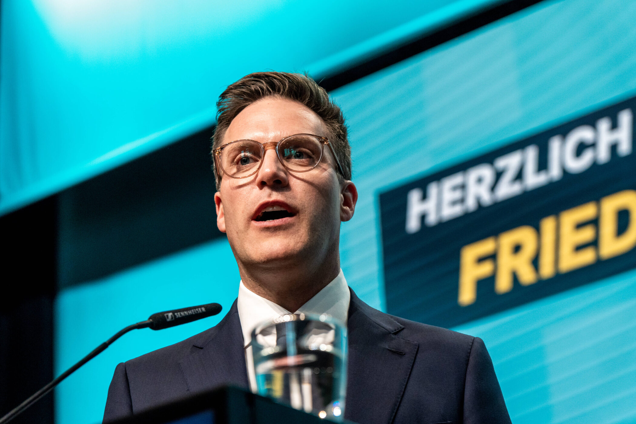 Mann mit Brille spricht am Mikrofon, Text "HERZLICH" im Hintergrund.