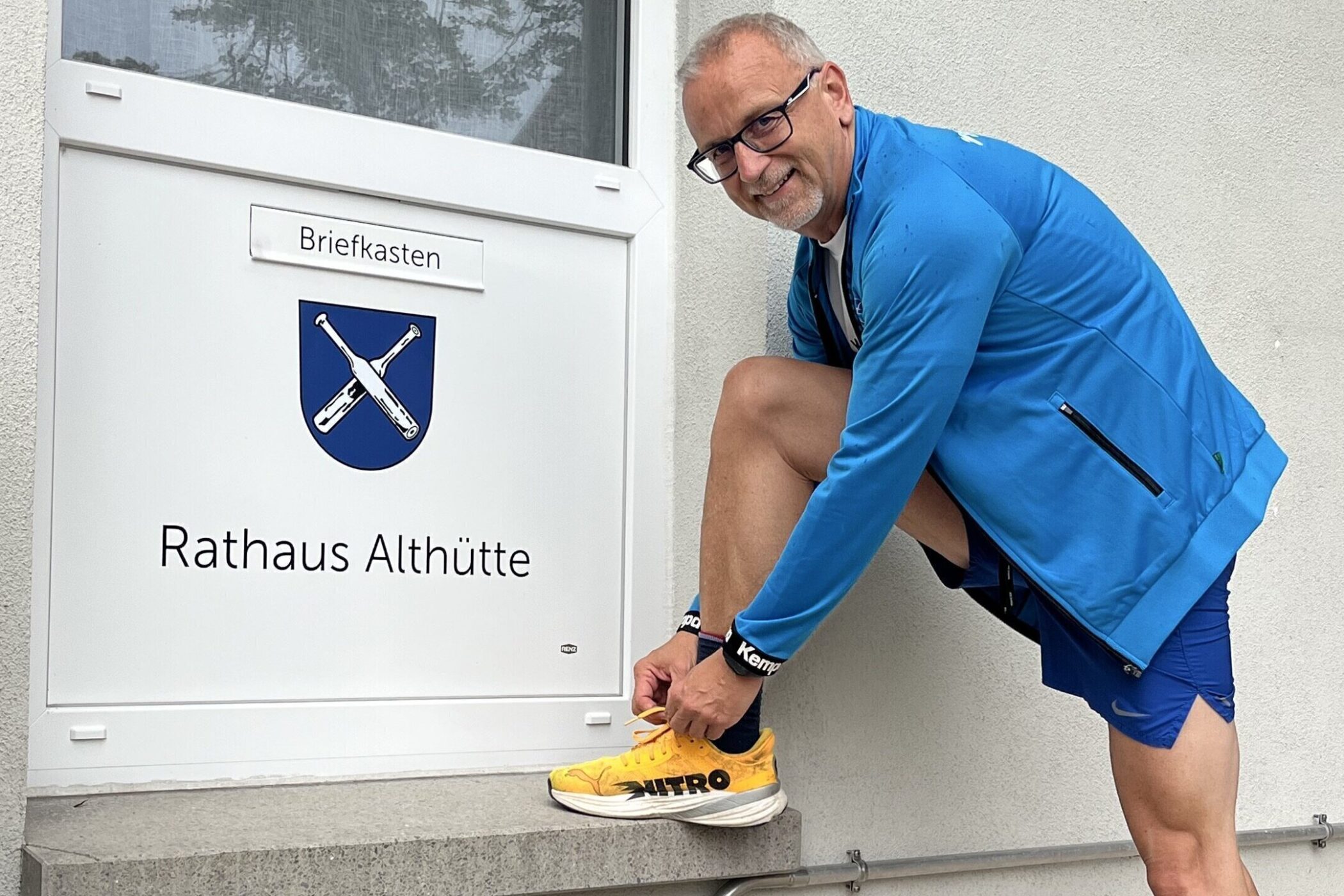 Mann bindet Schnürsenkel vor Schild "Rathaus Althütte" mit Wappen.