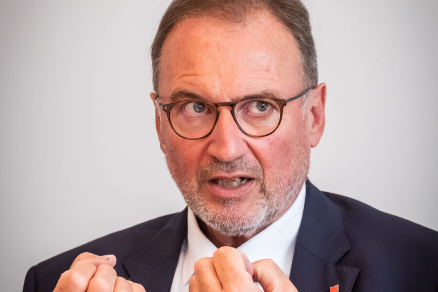 Mann mit Brille und Bart spricht gestikulierend, trägt Anzug und Krawatte.