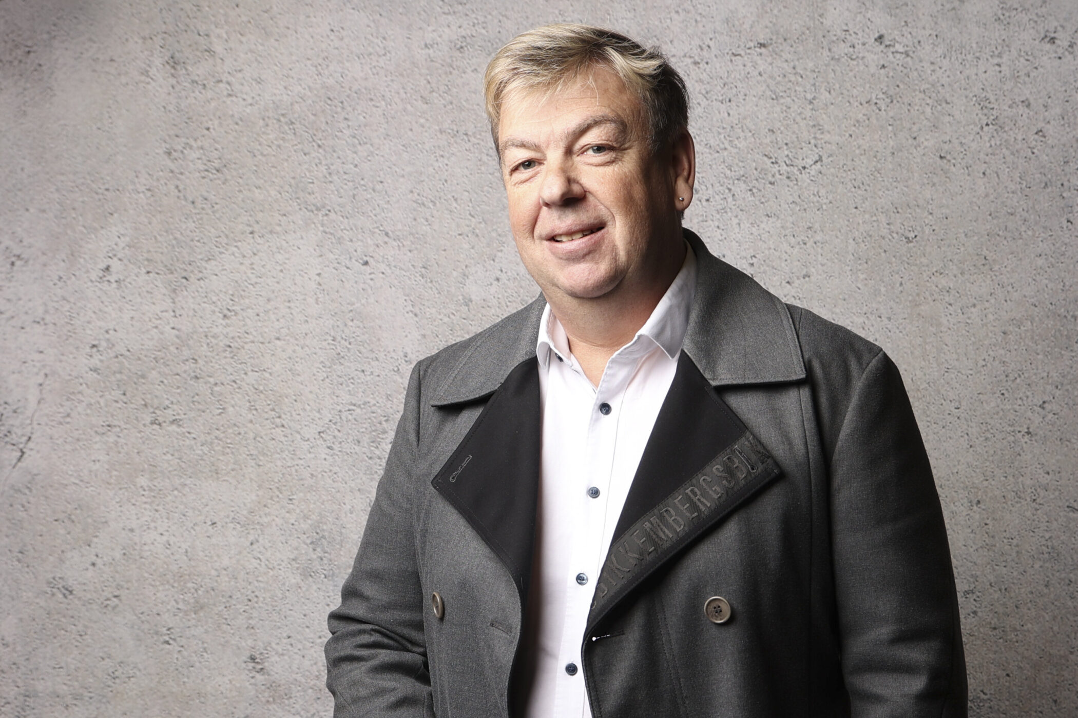 Mann in grauem Mantel und weißem Hemd vor grauer Wand.