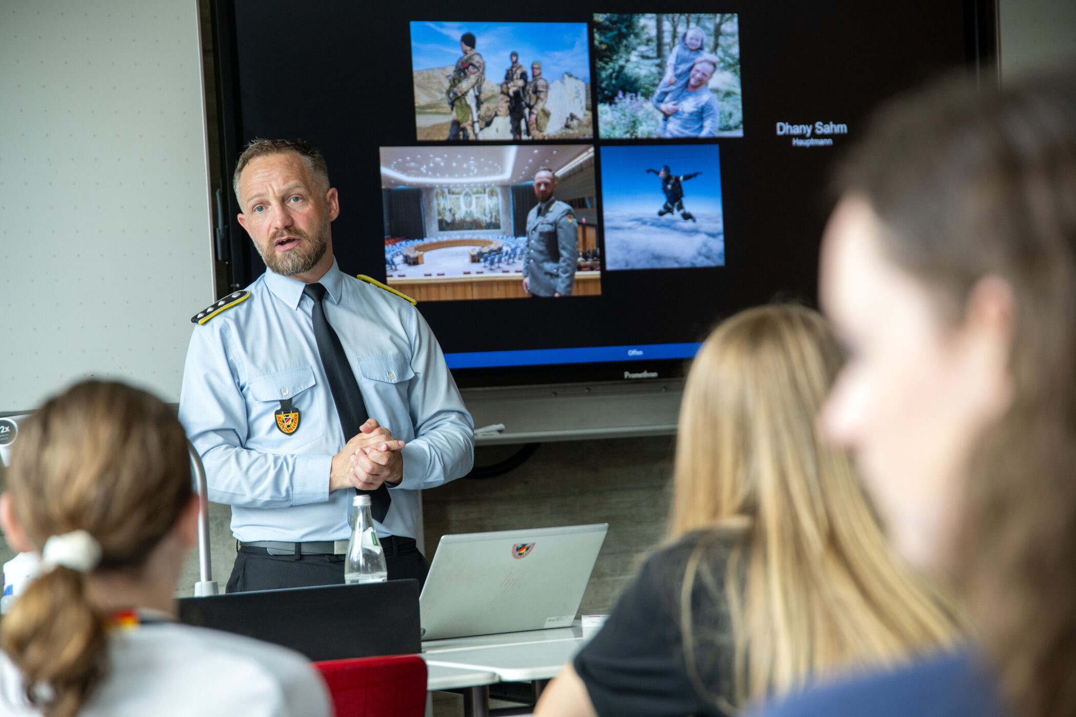 Mann in Uniform hält Vortrag vor Publikum, Bildschirm mit Bildern im Hintergrund.