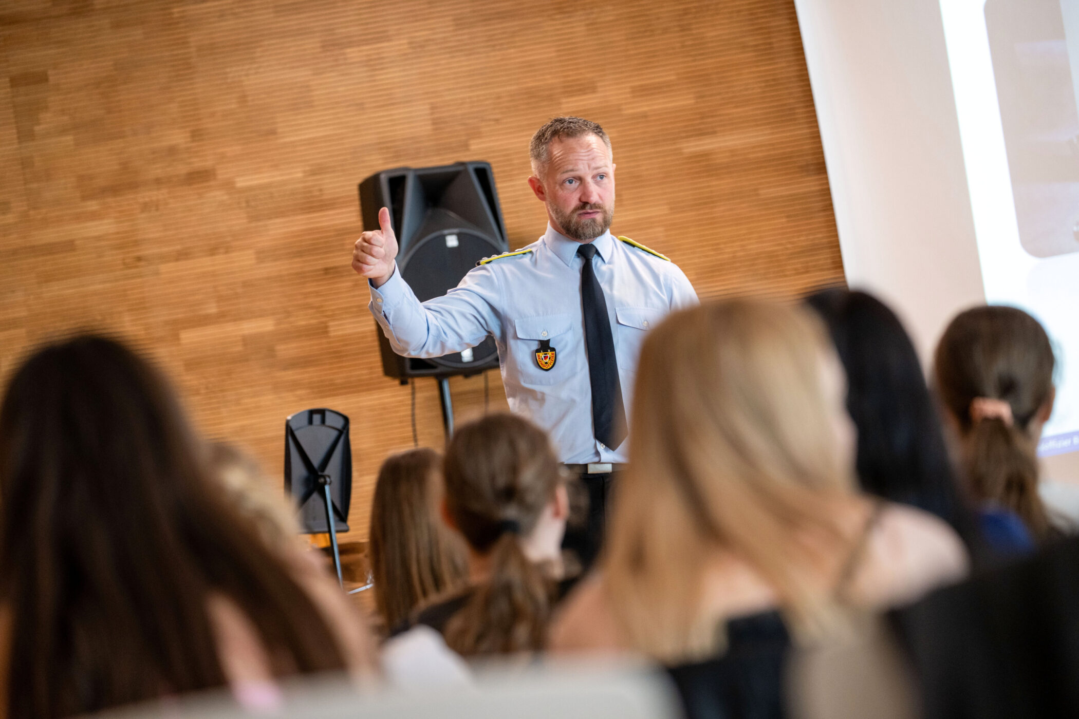 Mann in Uniform spricht vor Publikum, hebt Daumen, Holzwand im Hintergrund.