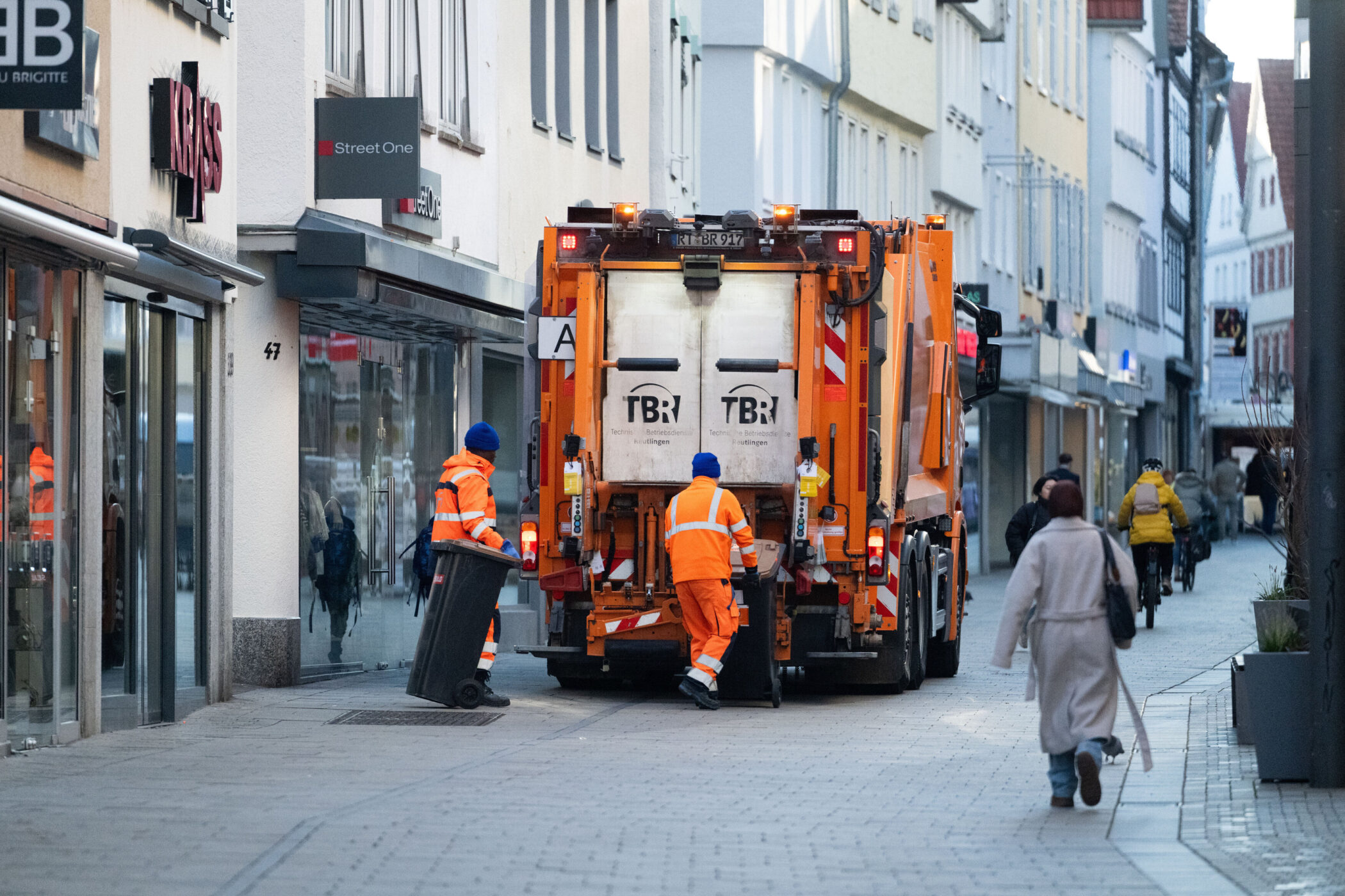 Zwei Müllmänner in orangefarbener Kleidung leeren Mülltonnen in ein Müllfahrzeug.