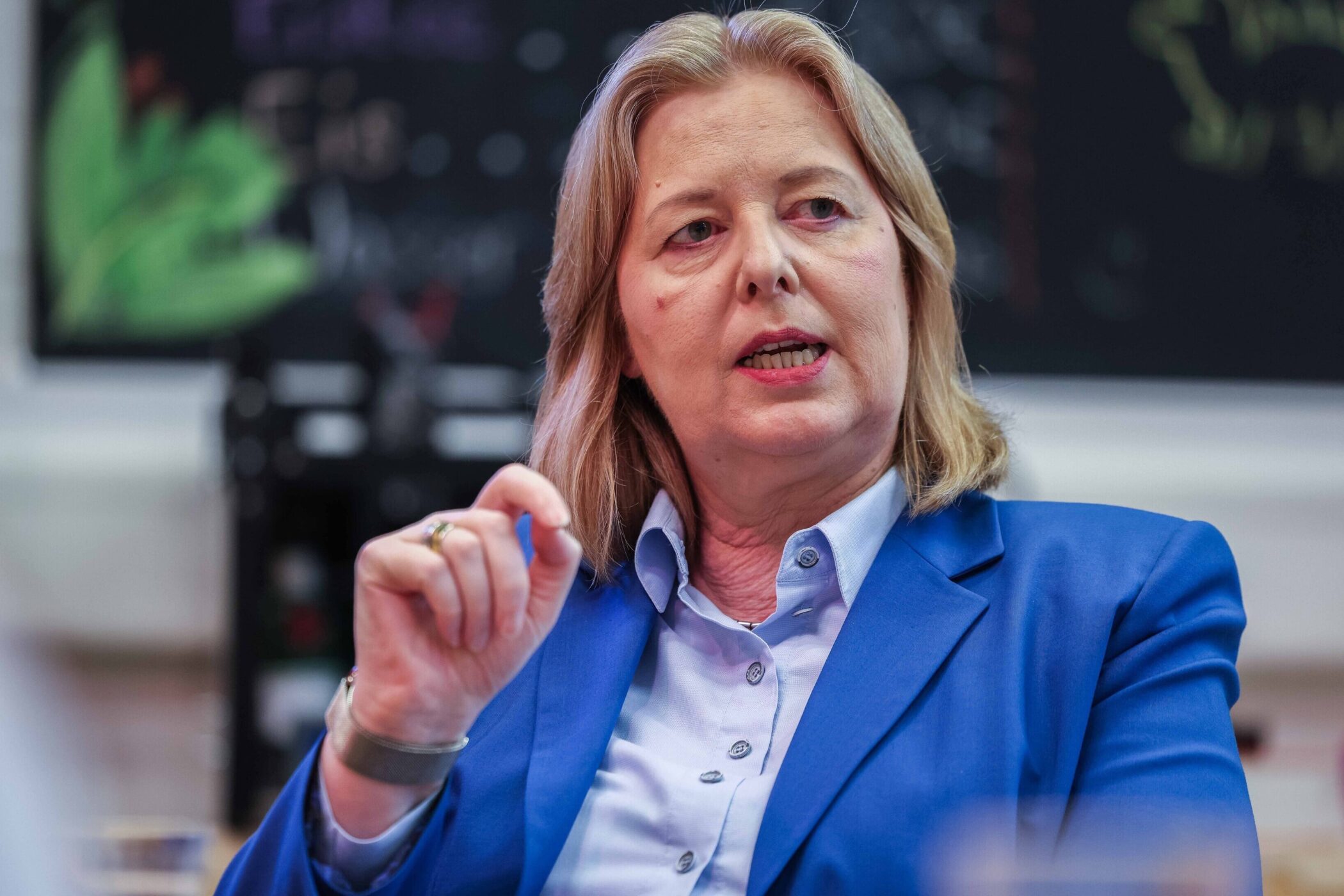 Eine Frau in blauem Blazer spricht gestikulierend. Hintergrund unscharf.