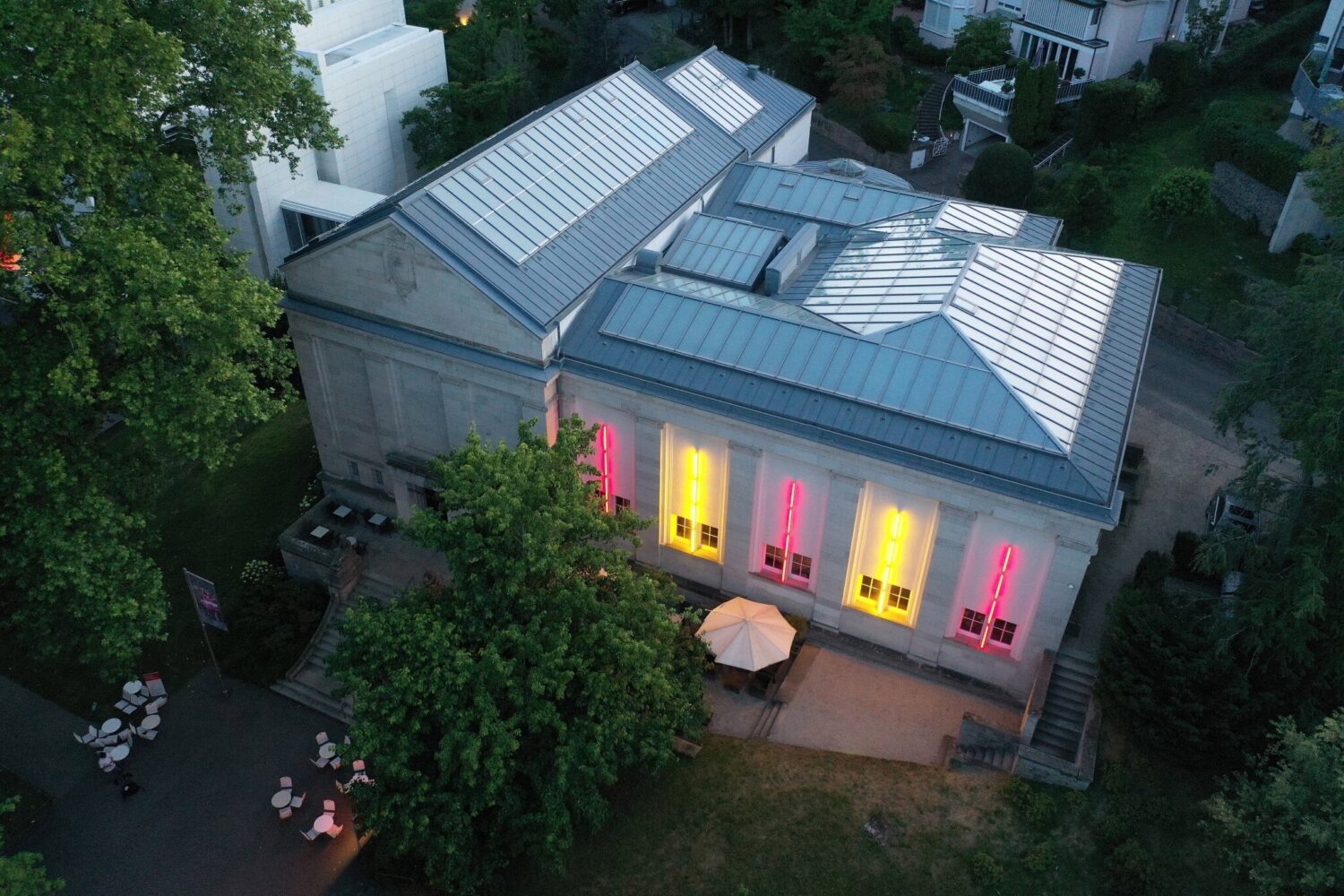 Gebäude mit beleuchteten Fenstern in Pink und Gelb, umgeben von Bäumen und Sitzbereich.
