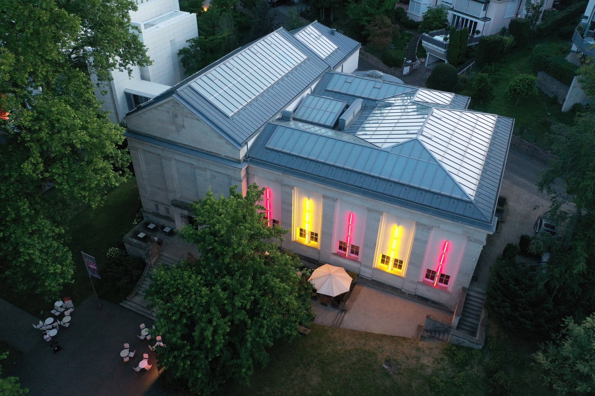 Gebäude mit beleuchteten Fenstern in Pink und Gelb, umgeben von Bäumen und Sitzbereich.