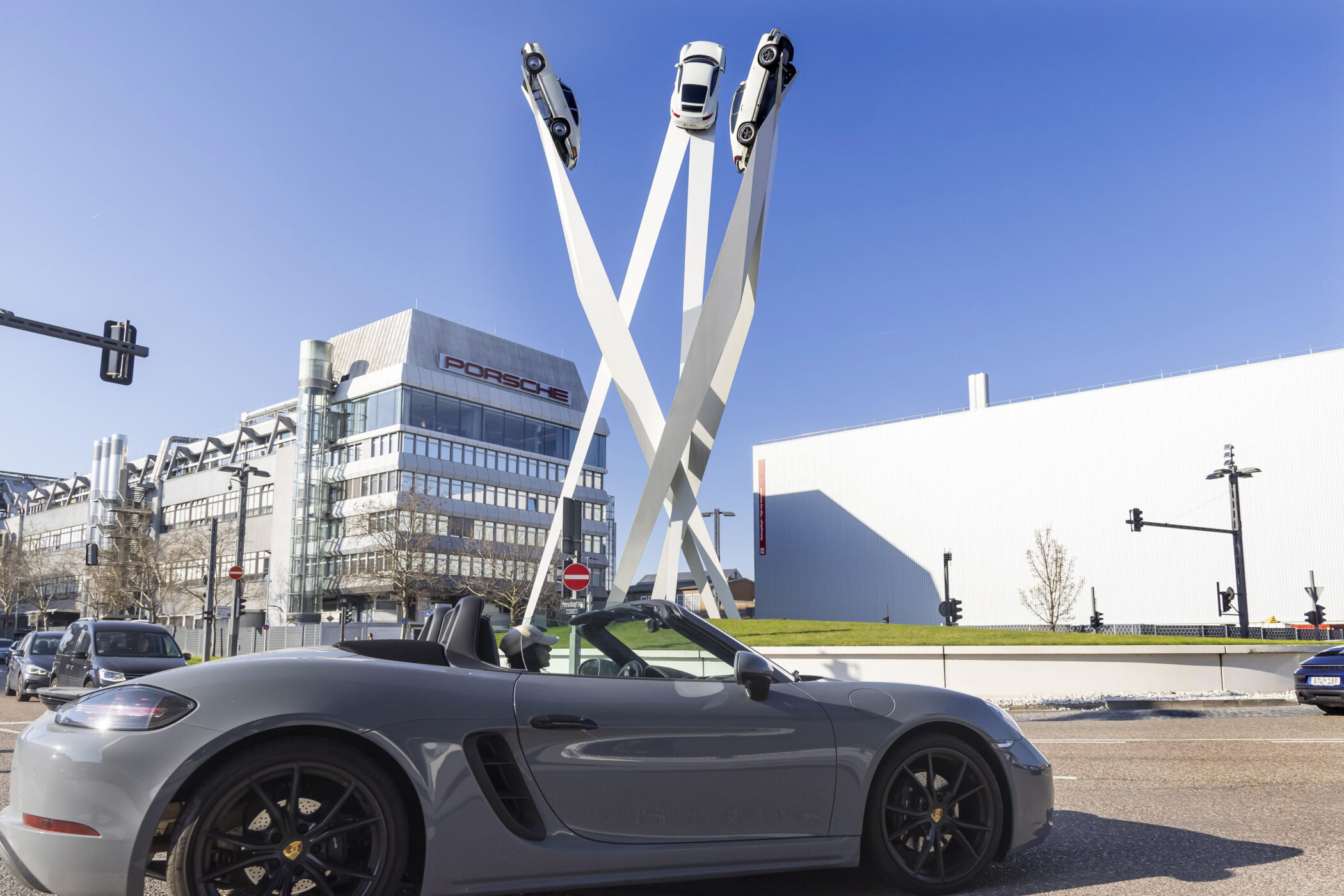 Graues Cabrio vor Porsche-Gebäude und Skulptur mit drei Autos auf Säulen.