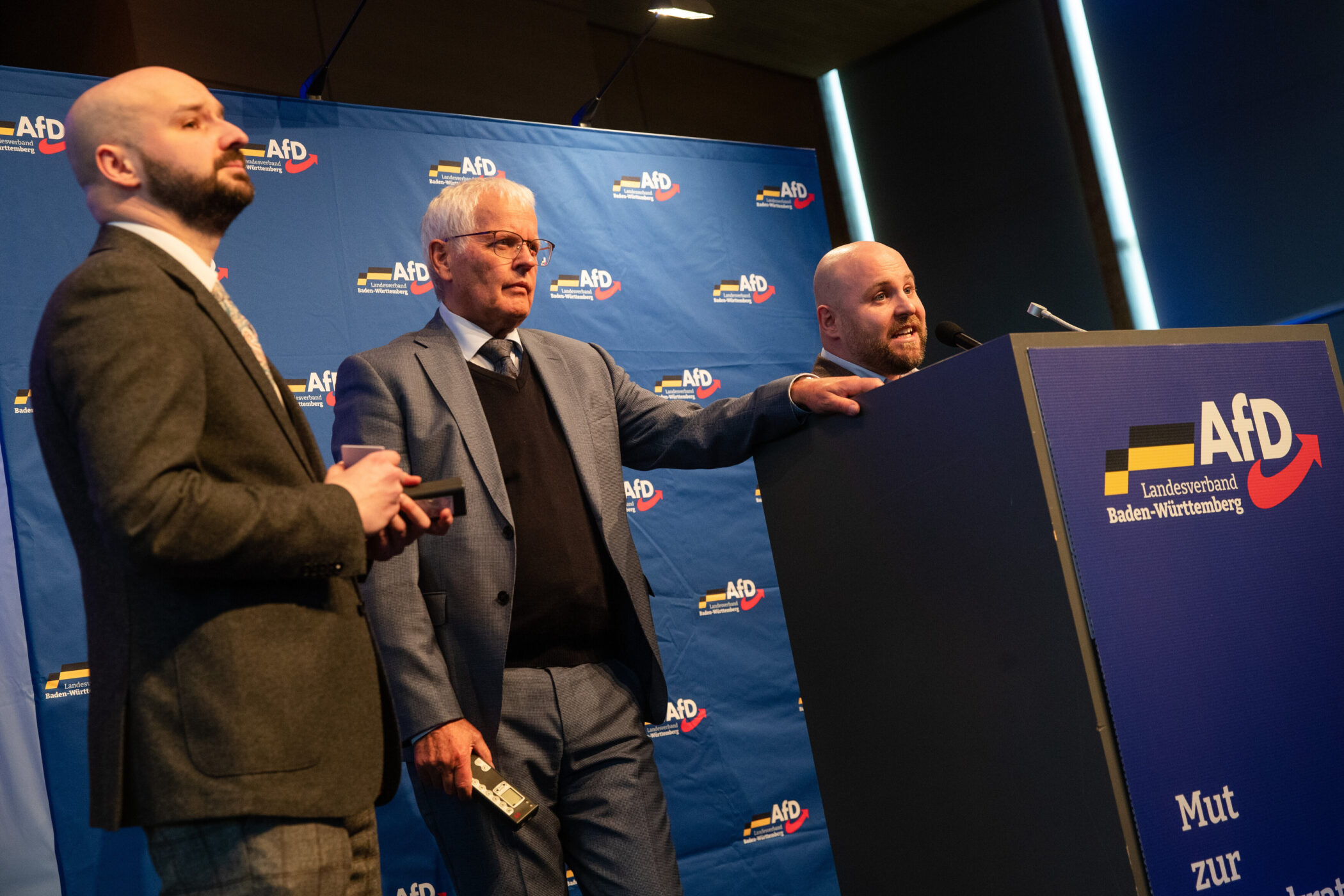 Drei Männer vor AfD-Logo, einer spricht am Rednerpult.