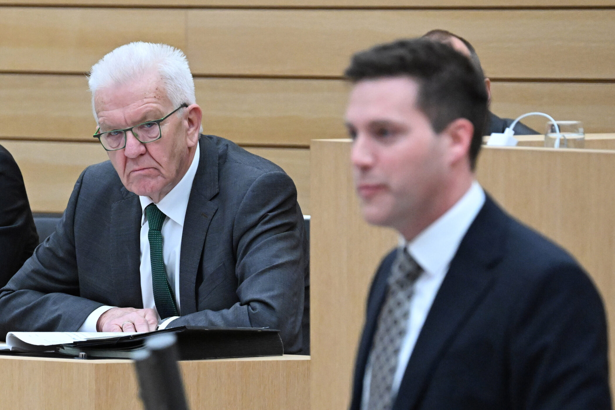 Zwei Männer in Anzügen in einem Sitzungssaal, einer spricht, der andere sitzt.