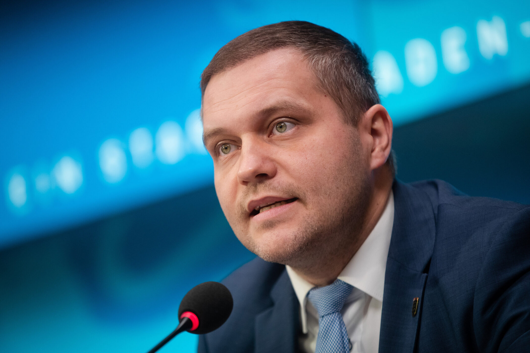 Mann im Anzug spricht in ein Mikrofon, blauer Hintergrund.