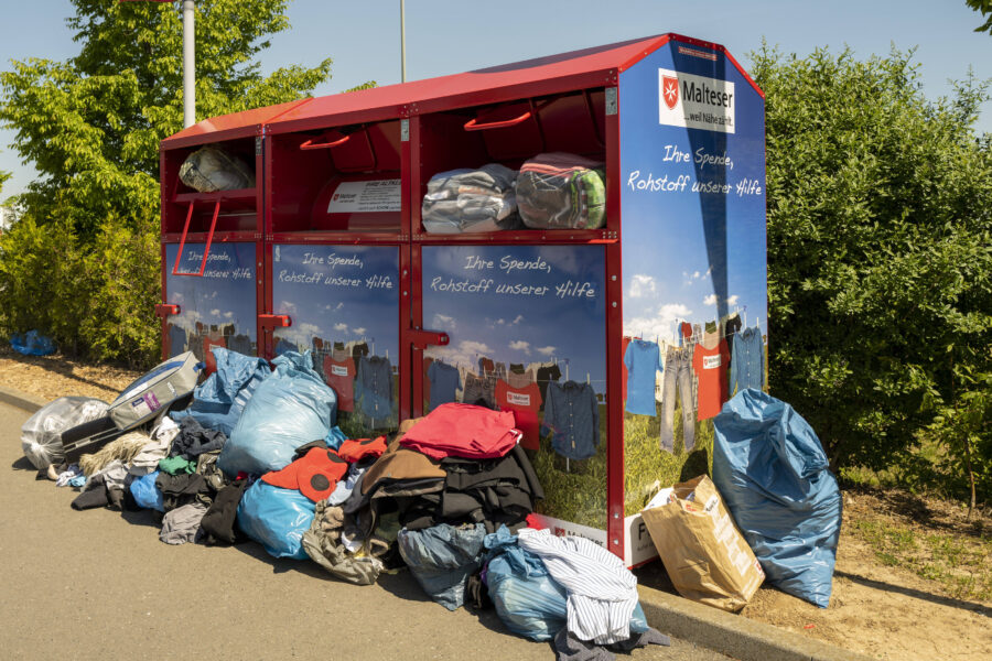 Rote Altkleidercontainer mit Kleidung und Säcken davor. Text: "Ihre Spende, Rohstoff unserer Hilfe".