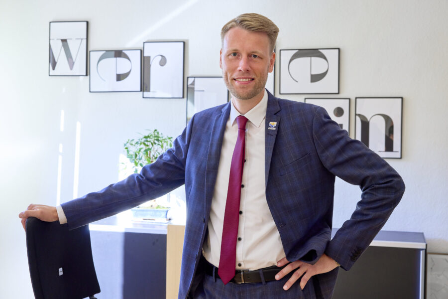 Mann im Anzug steht in Büro, lehnt an Stuhl, Bilder an der Wand.