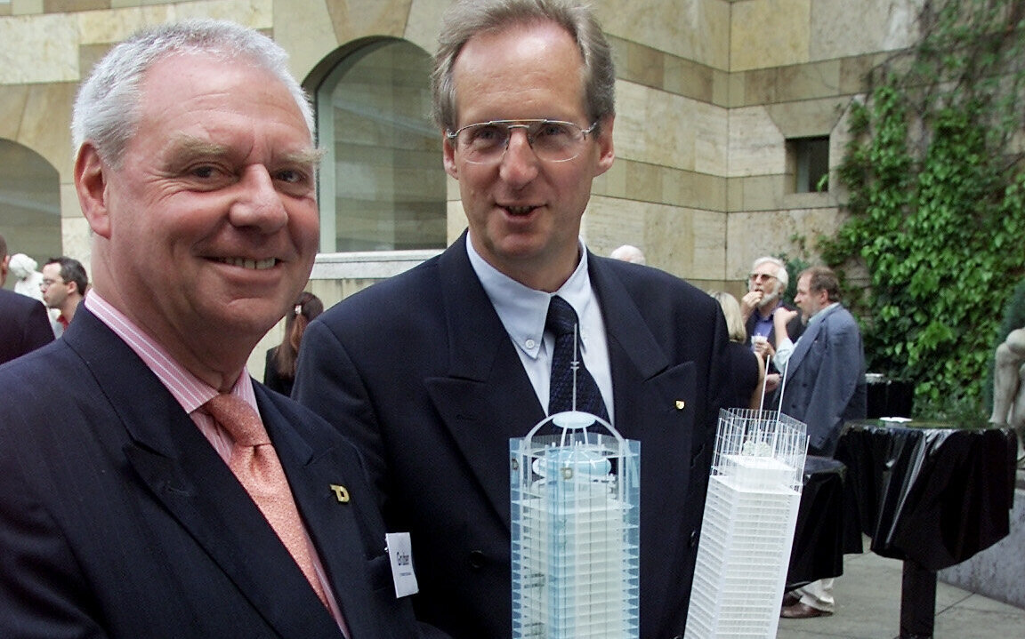Zwei Männer in Anzügen halten Architekturmodelle, im Hintergrund Menschen bei einer Veranstaltung.