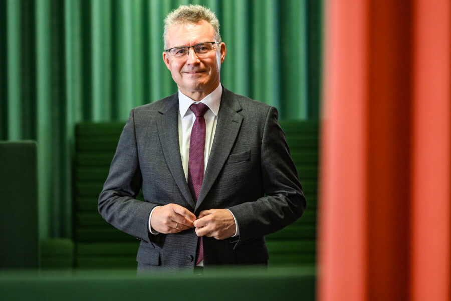 Ein Mann im Anzug steht vor grünen Vorhängen und lächelt in die Kamera.