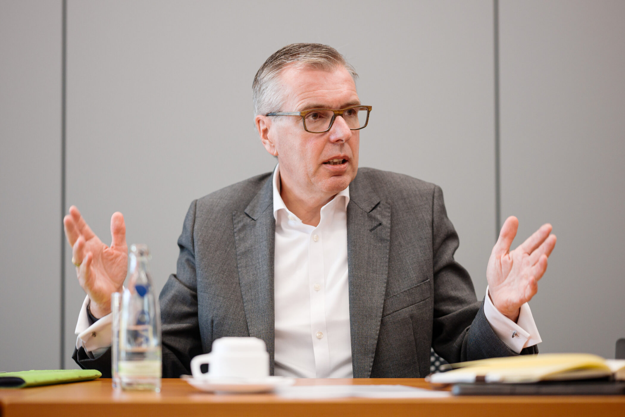 Mann in Anzug gestikuliert, sitzt an Tisch mit Wasserflasche und Tasse.