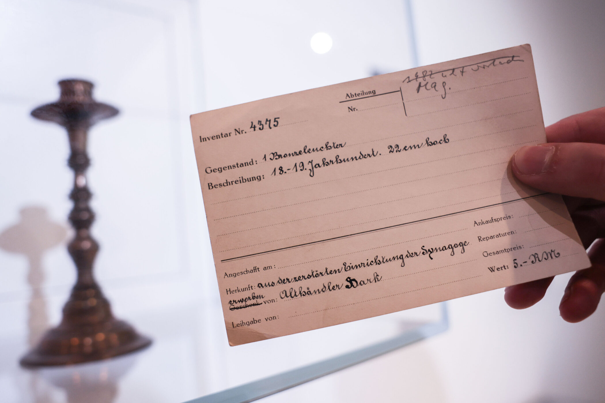 Hand hält Karte mit Inventarnummer, daneben unscharfer Kerzenhalter.