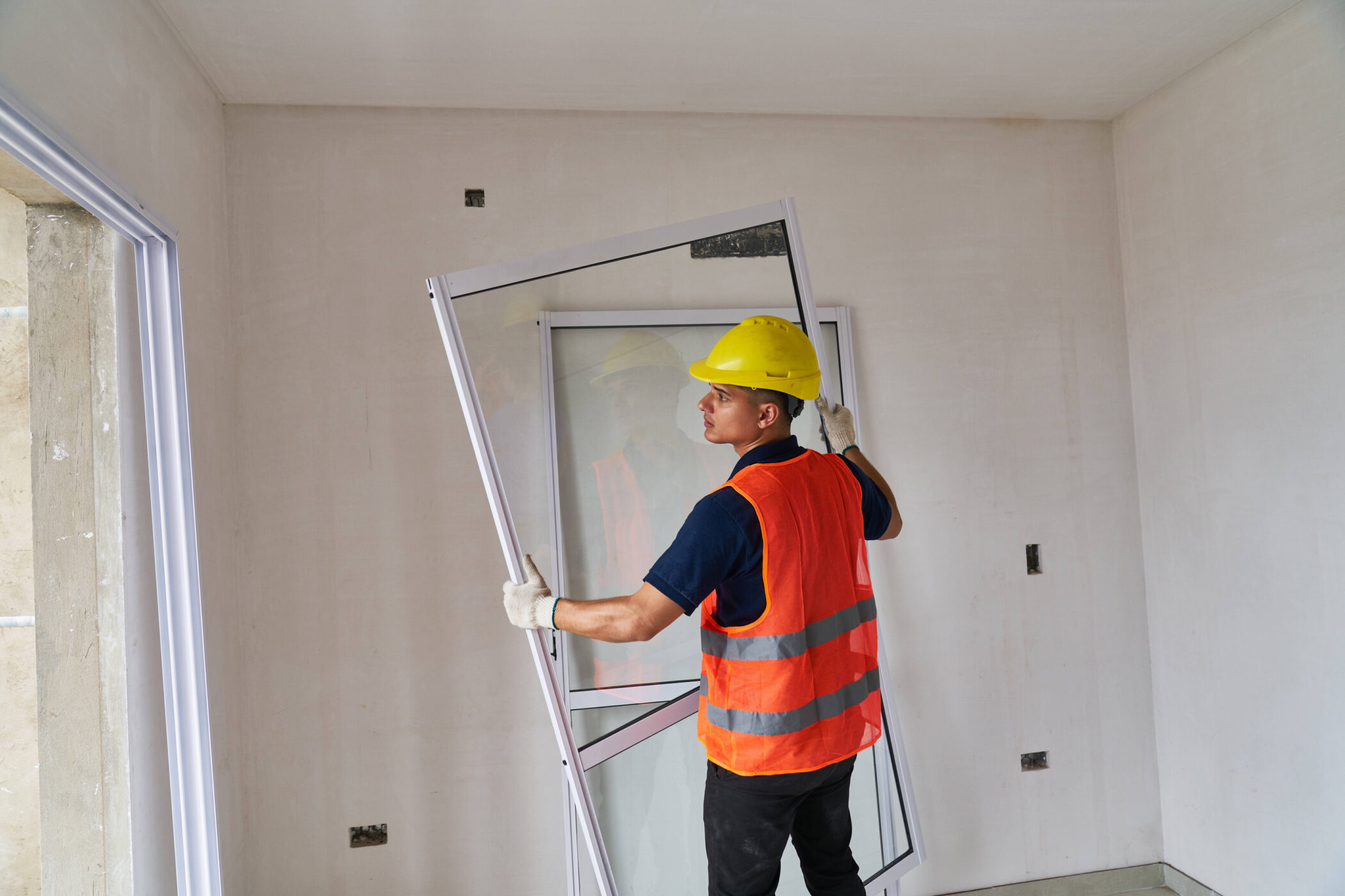 Arbeiter mit Helm und Weste montiert Fensterrahmen in einem Raum.