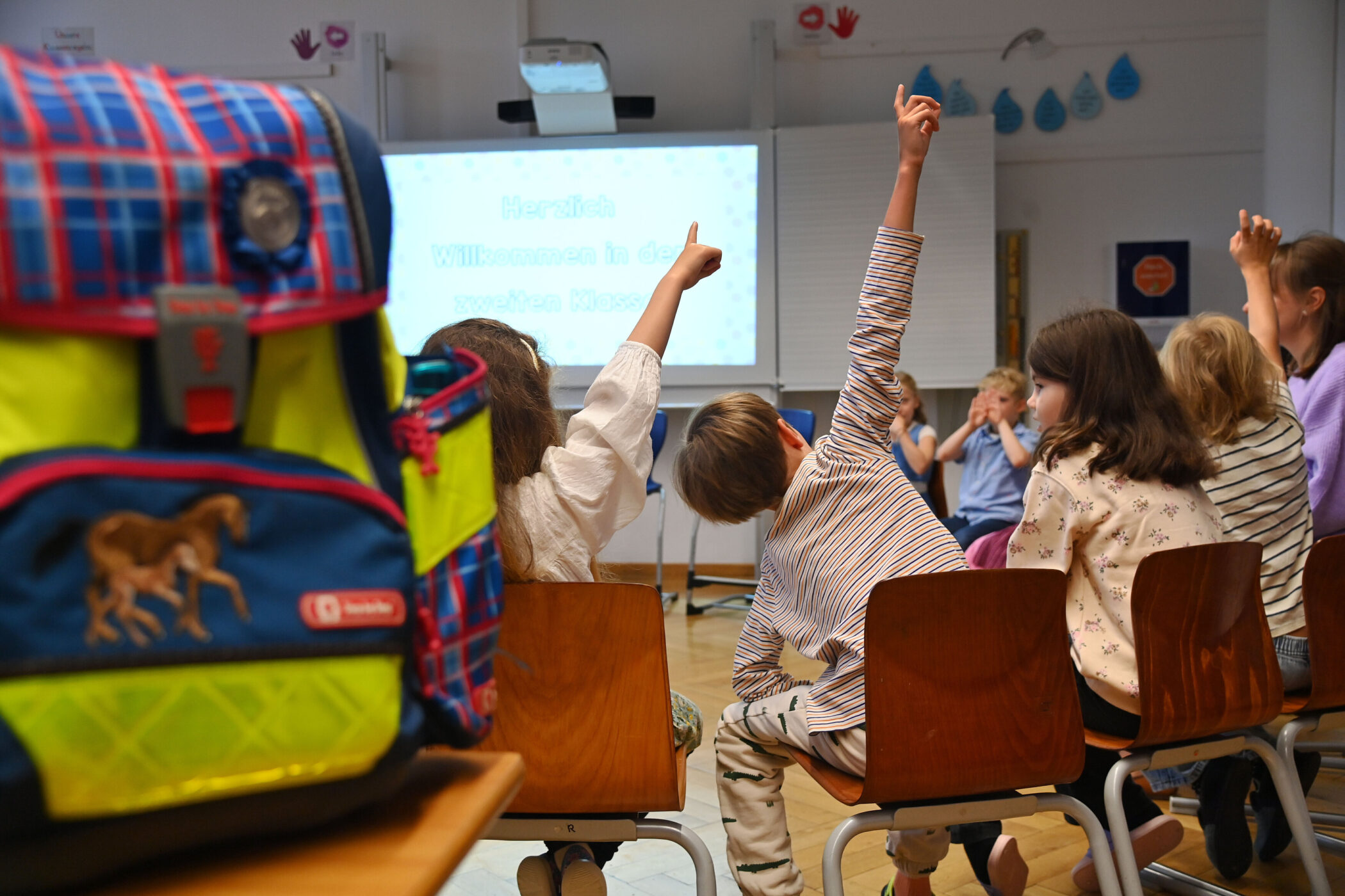 Kinder sitzen auf Stühlen, einige heben die Hand. Ein bunter Rucksack im Vordergrund.