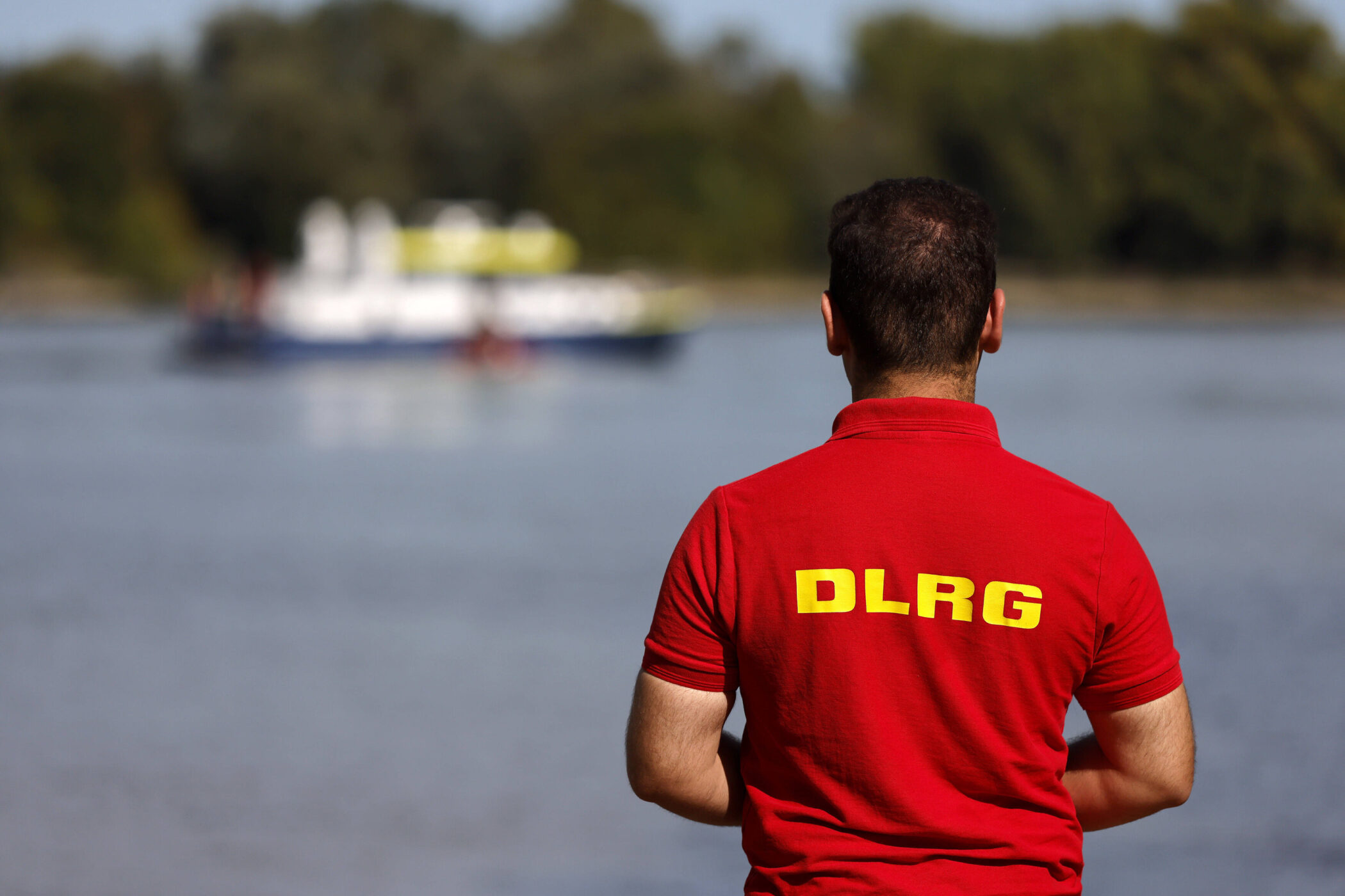 Person in rotem DLRG-Shirt blickt auf einen Fluss, im Hintergrund ein unscharfes Boot.