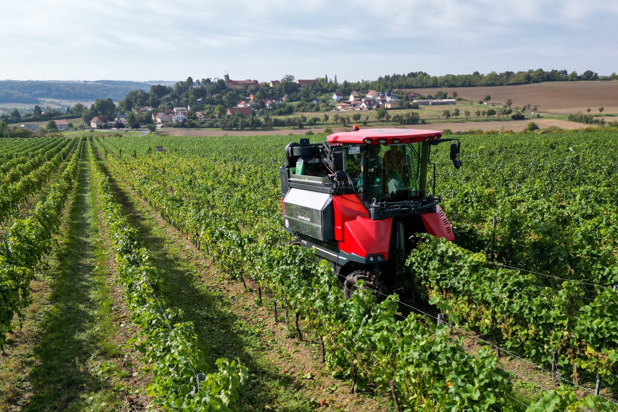 Roter Traktor fährt durch Weinreben, im Hintergrund ein Dorf.