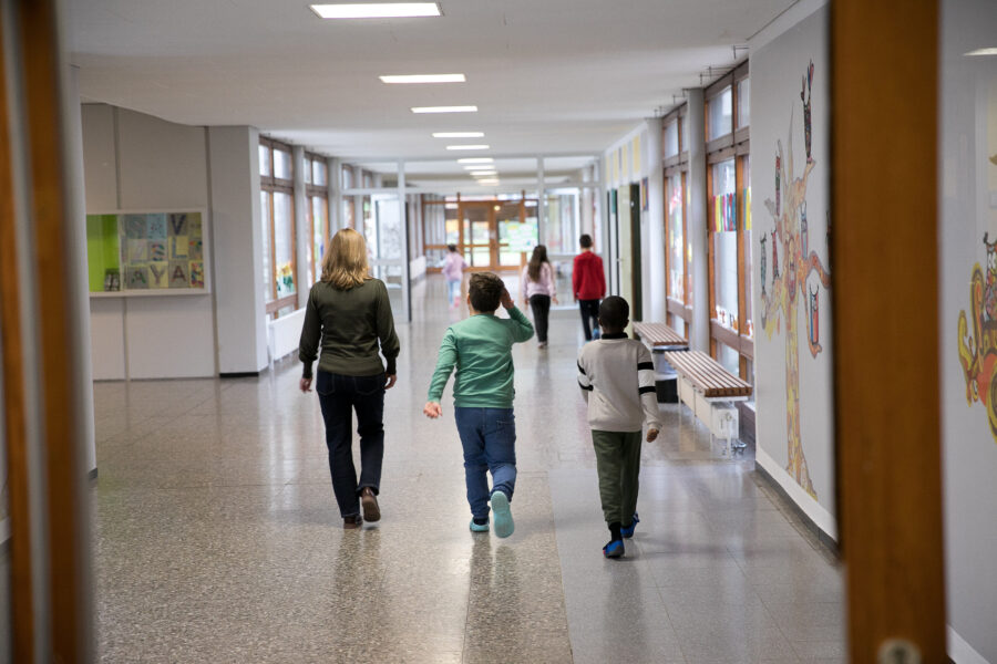 Person und zwei Kinder gehen durch einen Schulflur, Wände mit Zeichnungen.