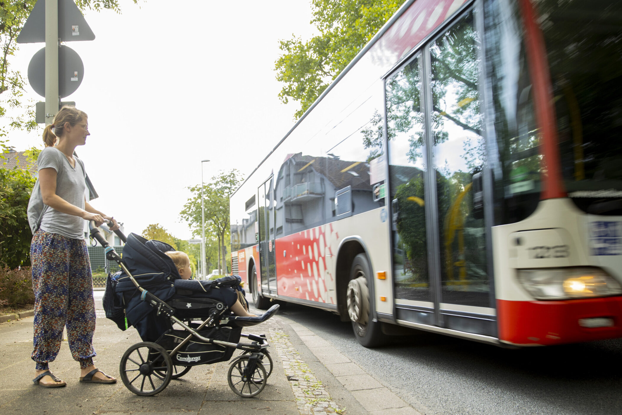 Eine Person mit Kinderwagen steht am Gehweg, ein Bus fährt vorbei.