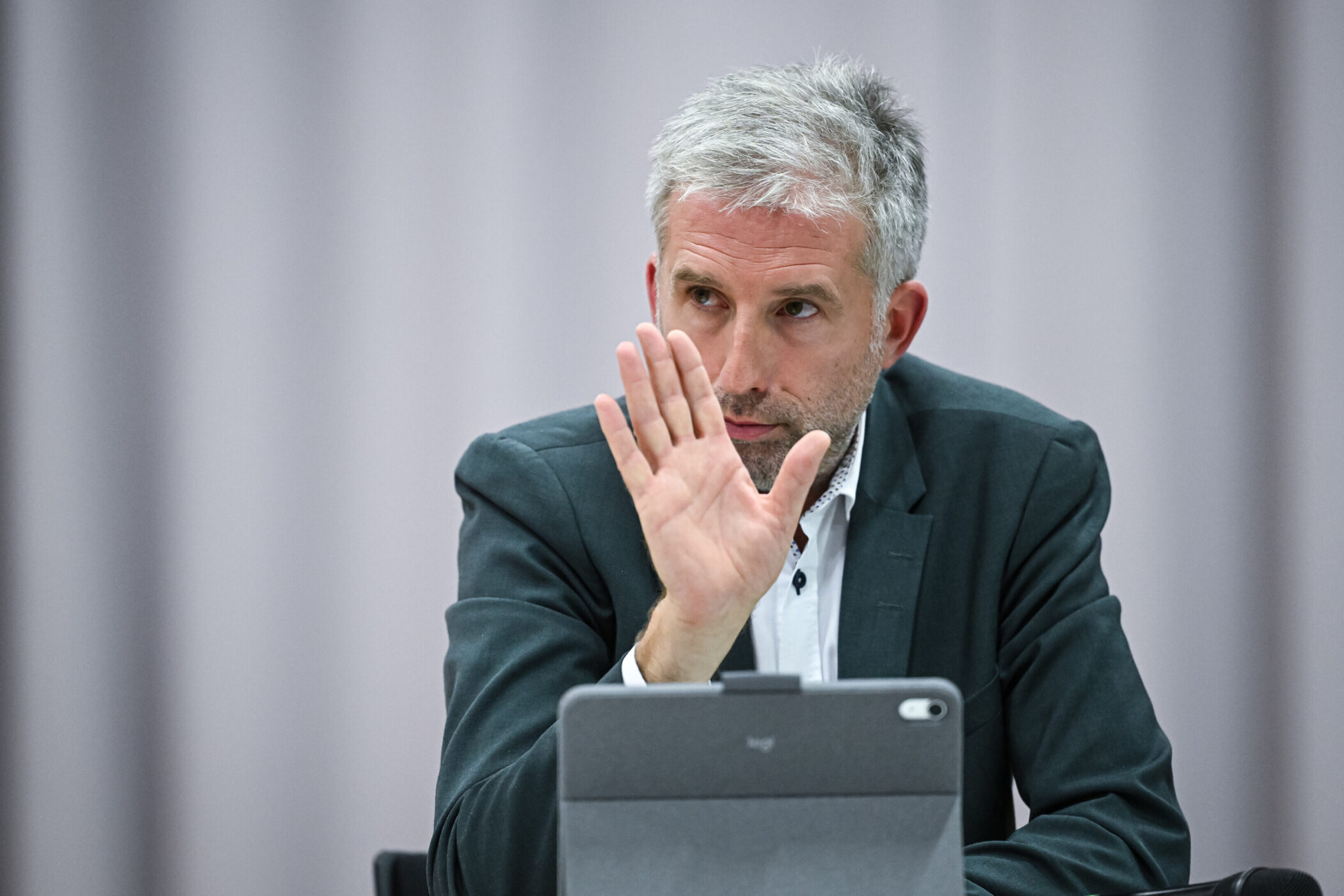 Mann in Anzug hebt Hand, vor einem Tablet sitzend.